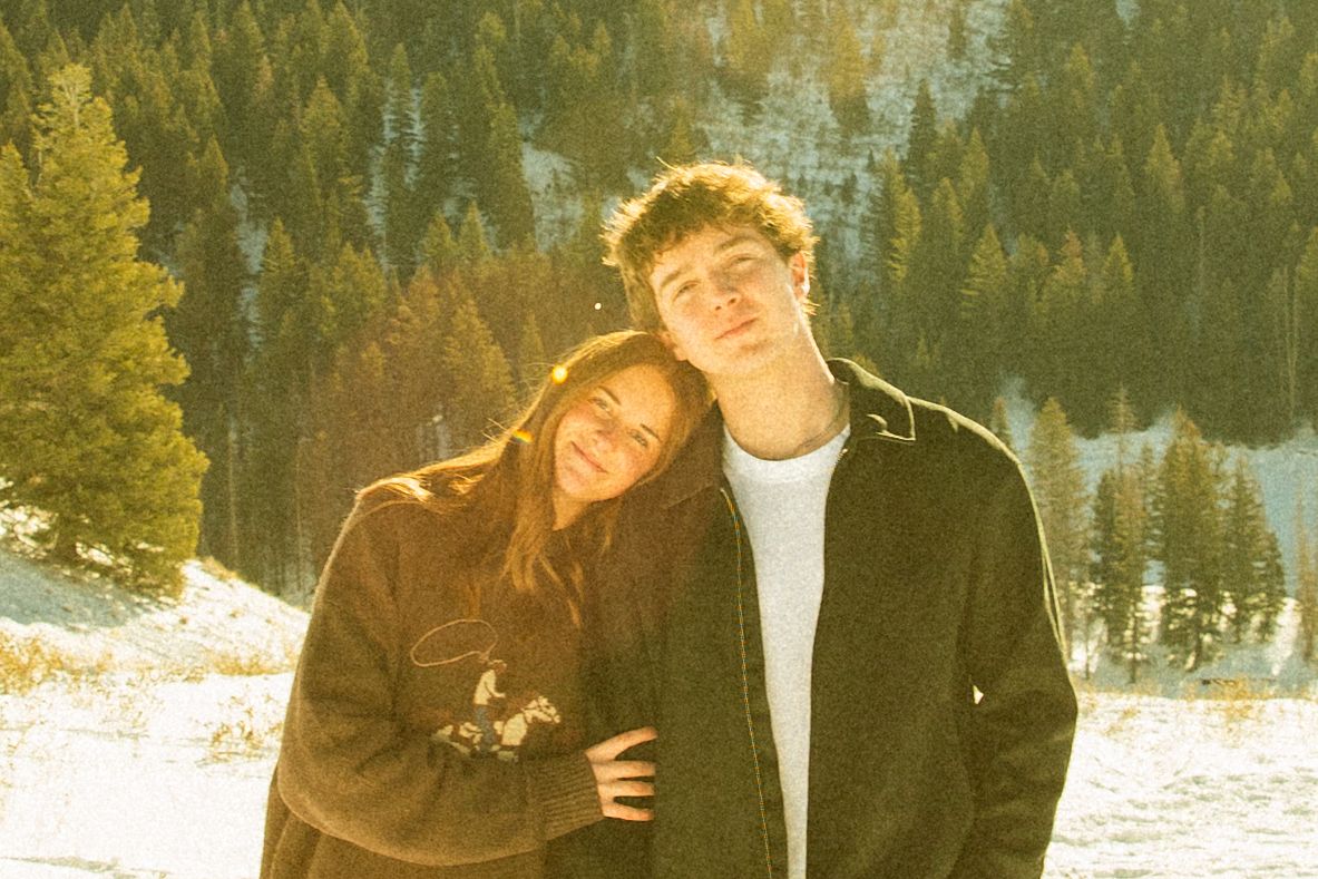 A couple poses together in a snowy winter landscape with evergreen trees in the background.