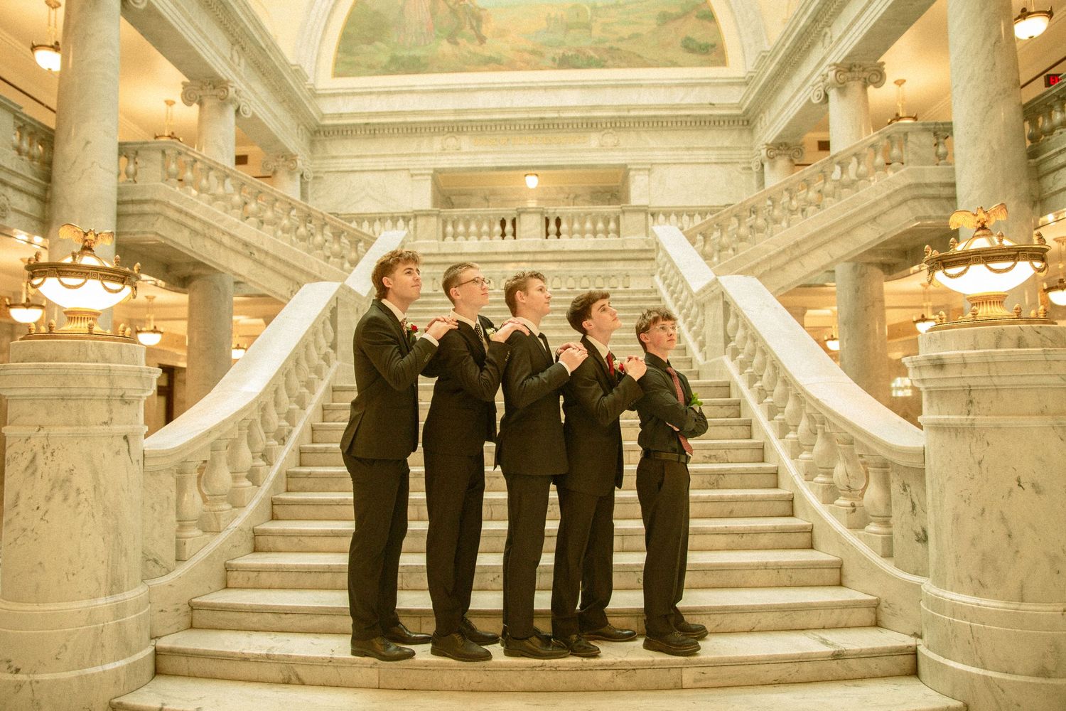 A group of performers in formal black attire stand on ornate marble stairs in a grand historic building interior.