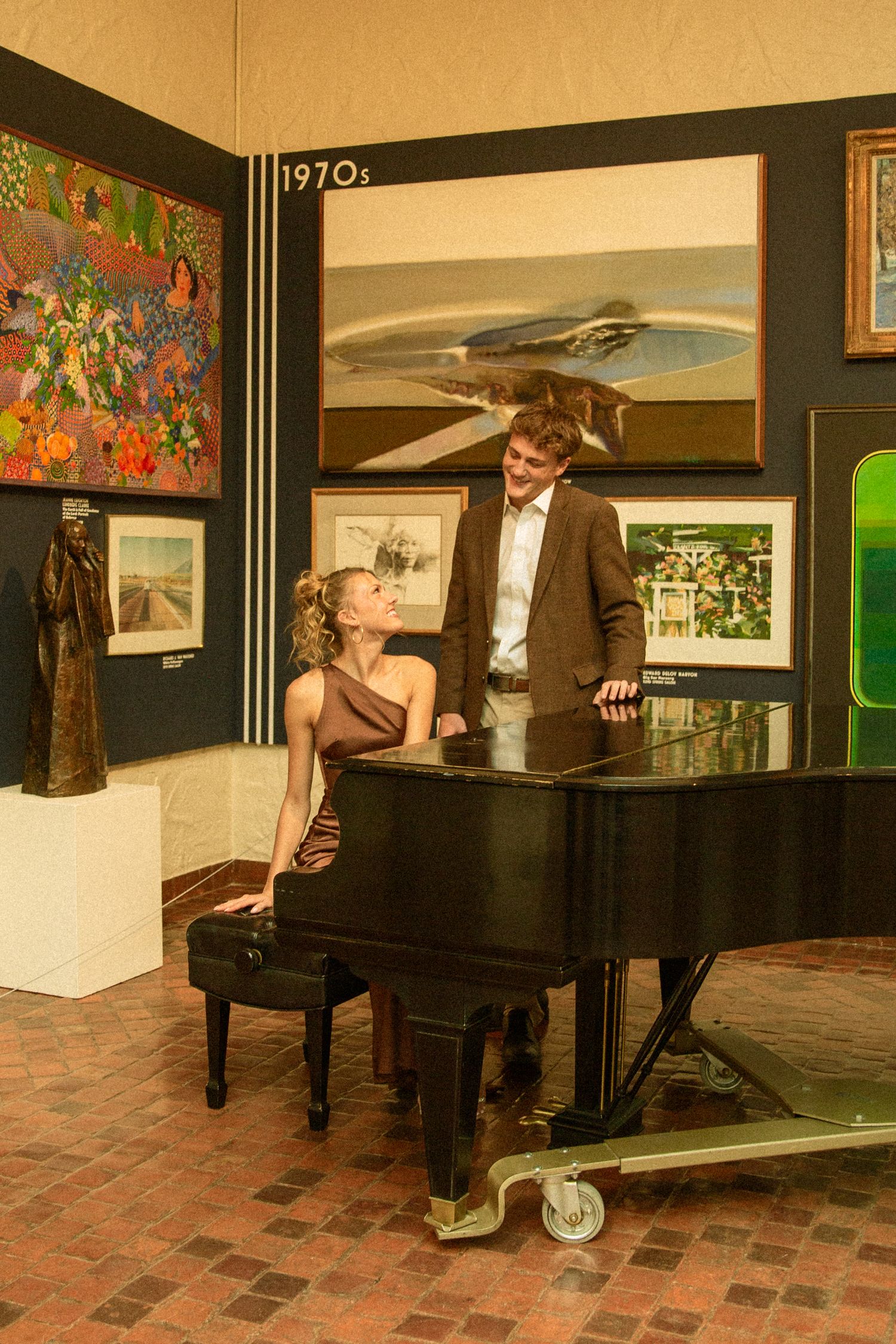 A pianist in formal attire performs at a black grand piano in a brick-floored room with artwork on the walls.