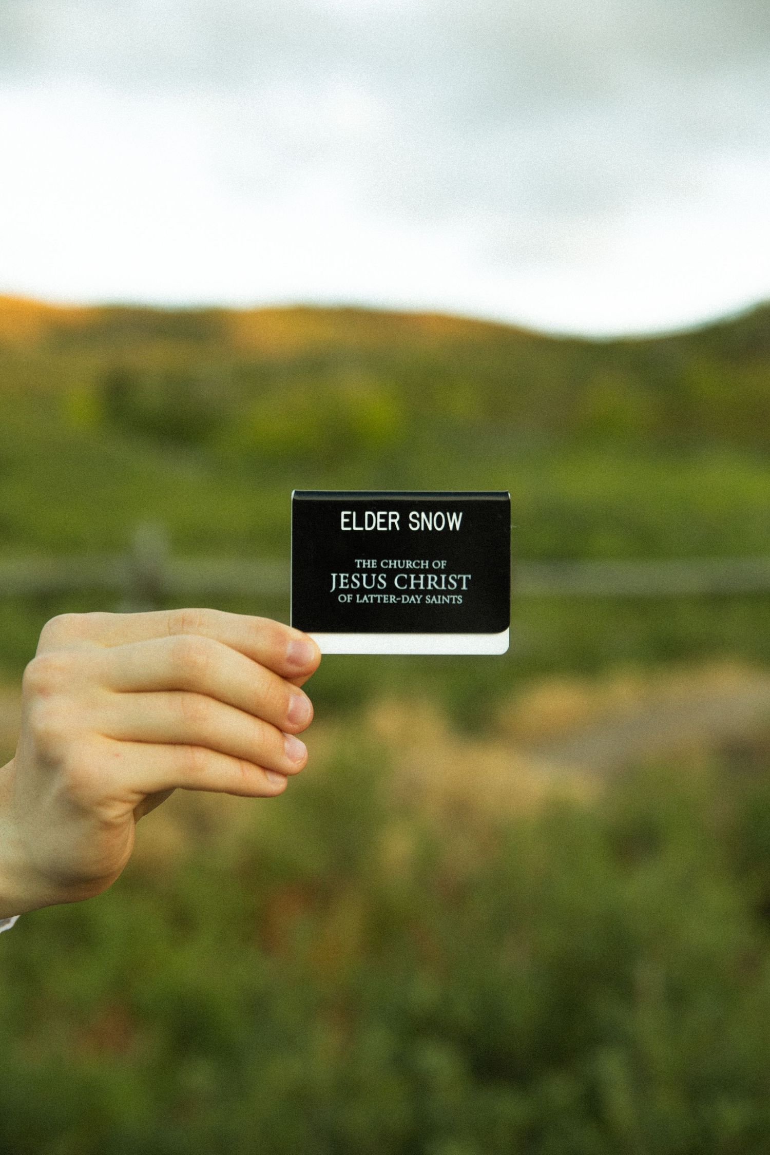 A hand holds up a black name tag against a blurred green natural background.