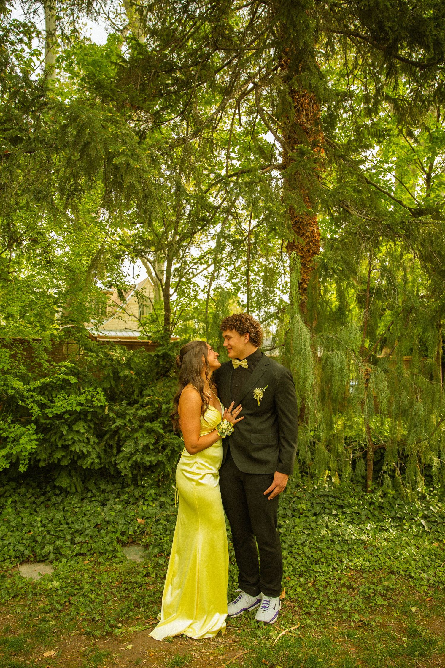 Couple in prom attire posing under a weeping tree, woman in yellow gown, man in black suit with bow tie.