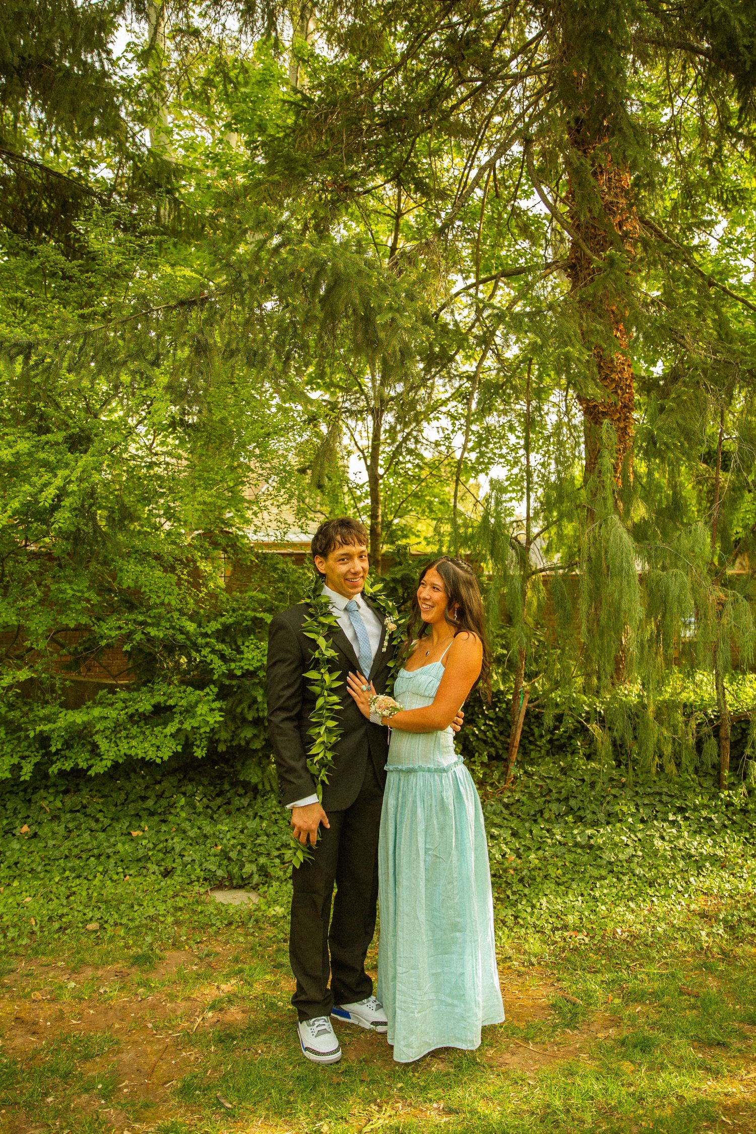 A young couple in formal attire poses outdoors under lush green trees, smiling together on a grassy lawn.