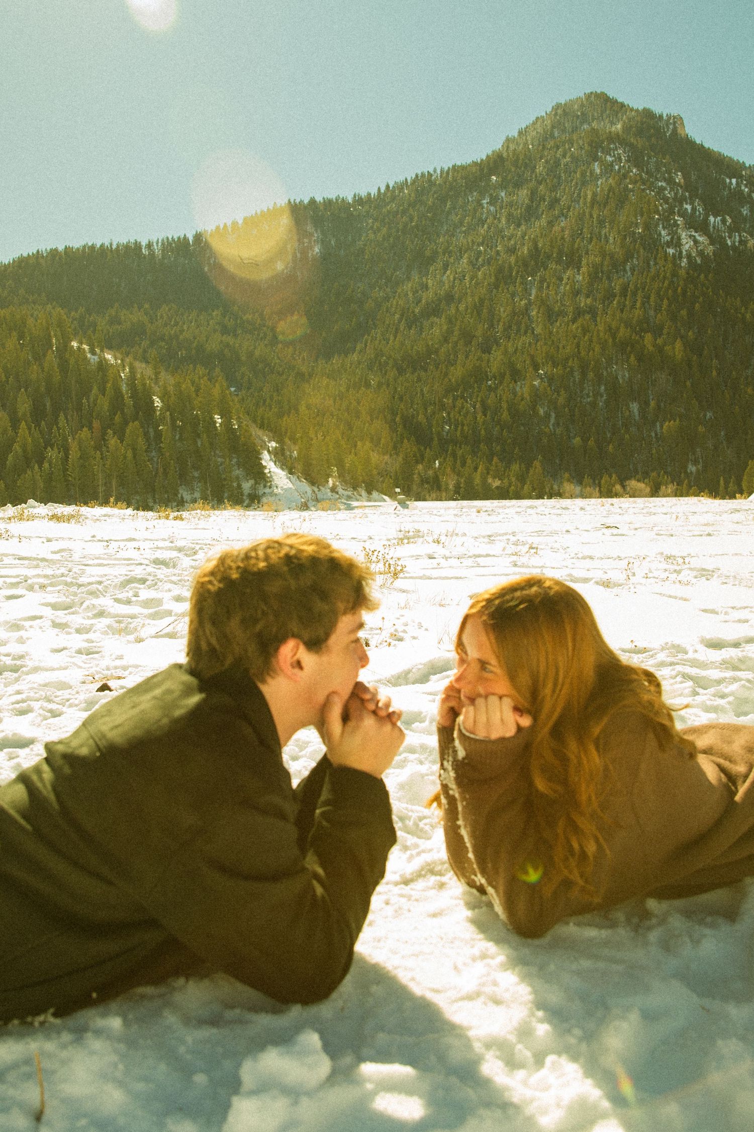 Two people lying in snow with mountains and forest in background during golden hour.