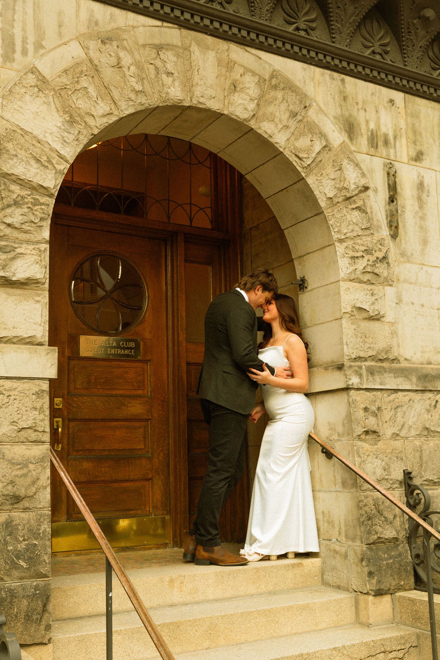 A couple shares a romantic moment under a stone archway entrance at a historic building.