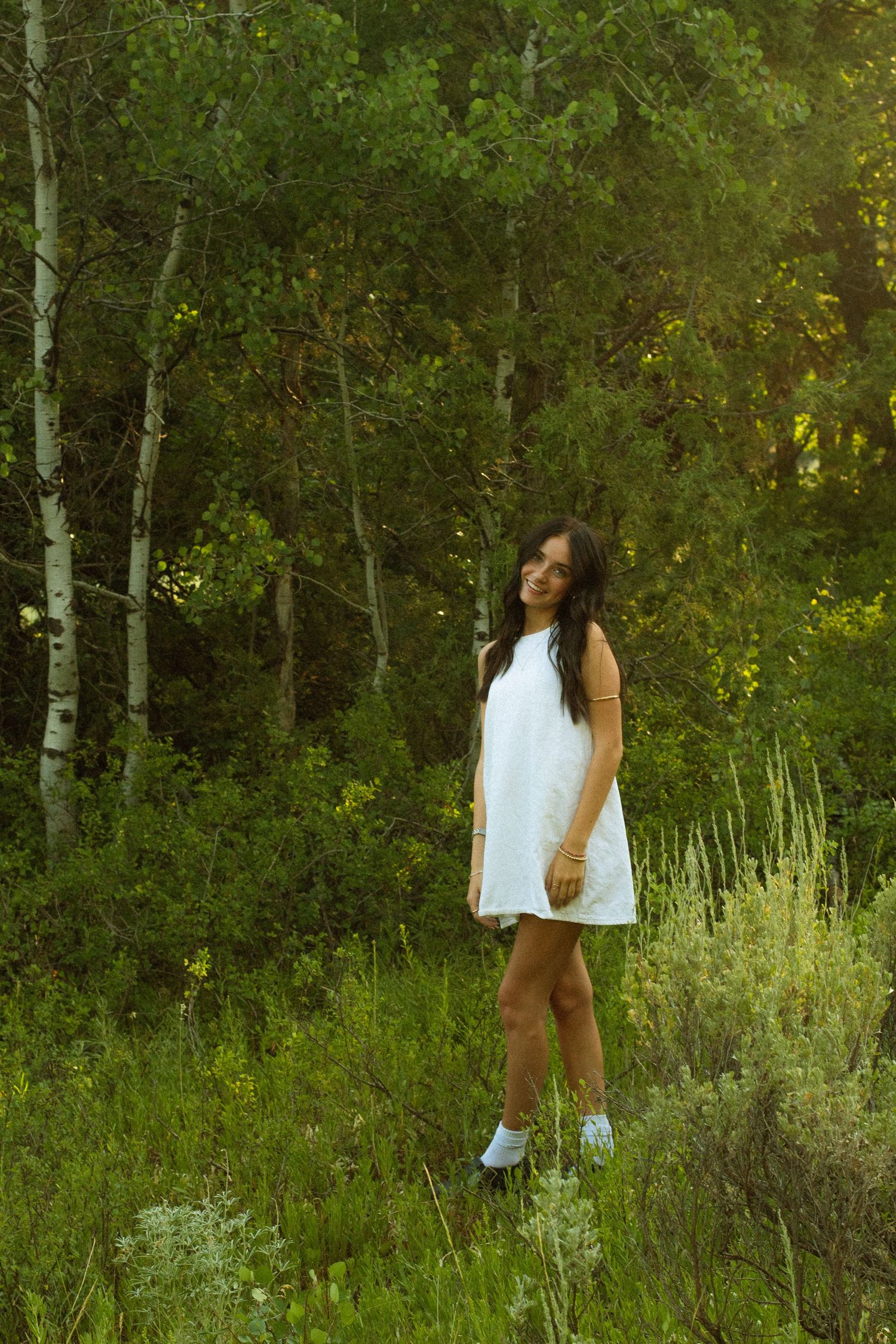 A person in a white top and shorts walks through a lush forest setting with green foliage and birch trees.