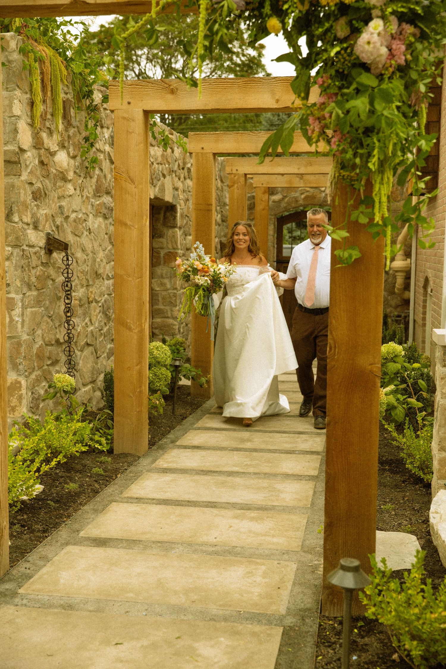 A stone-walled walkway decorated with wooden pergolas and greenery for a wedding ceremony.