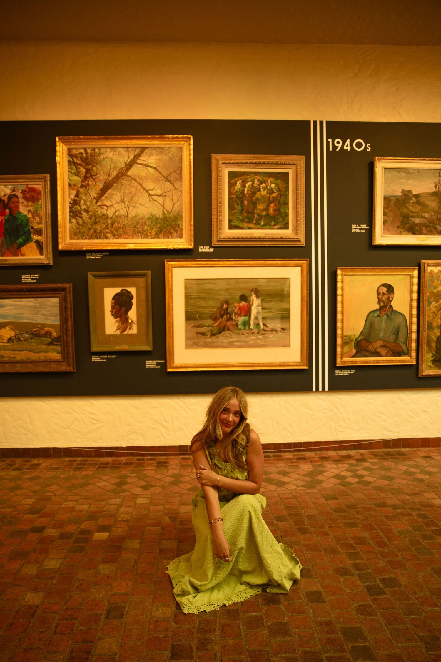 A young woman in a green dress sits on a tiled floor in front of a 1940s art gallery wall display.