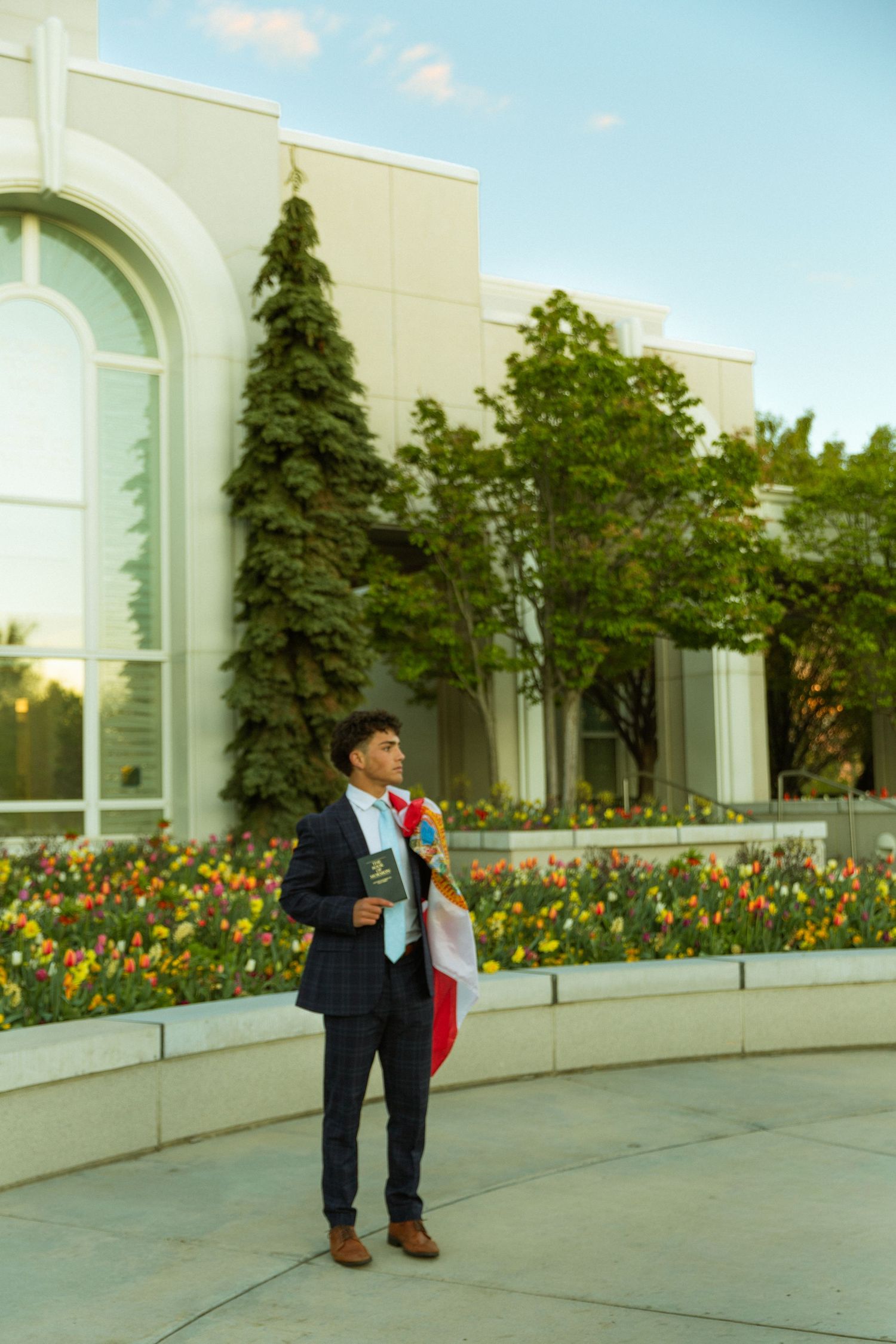 Young man in dark suit holding books and colorful sash stands outdoors near tulip garden and modern building.
