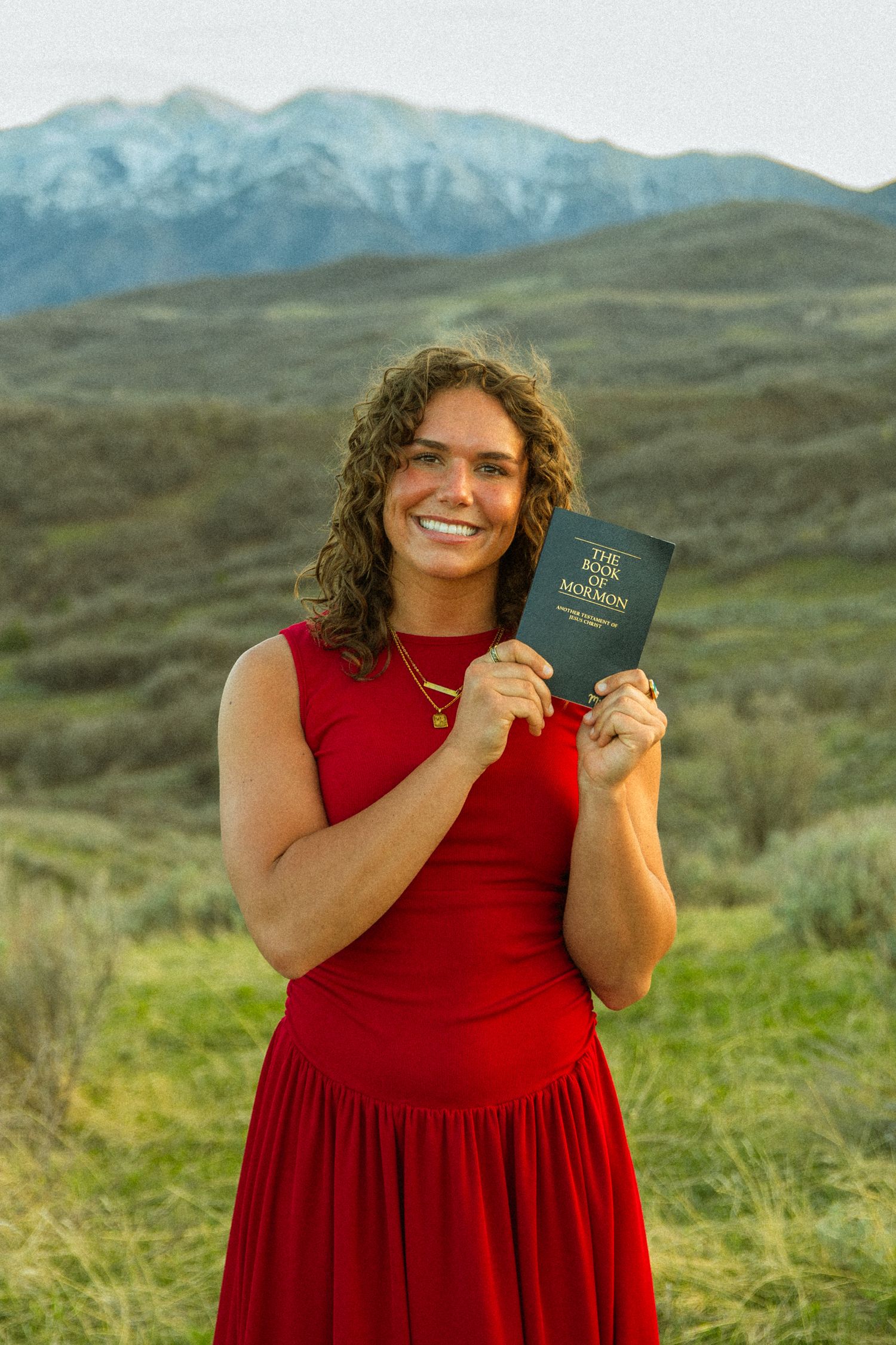 Smiling woman in red dress holding Book of Mormon outdoors with mountain backdrop.