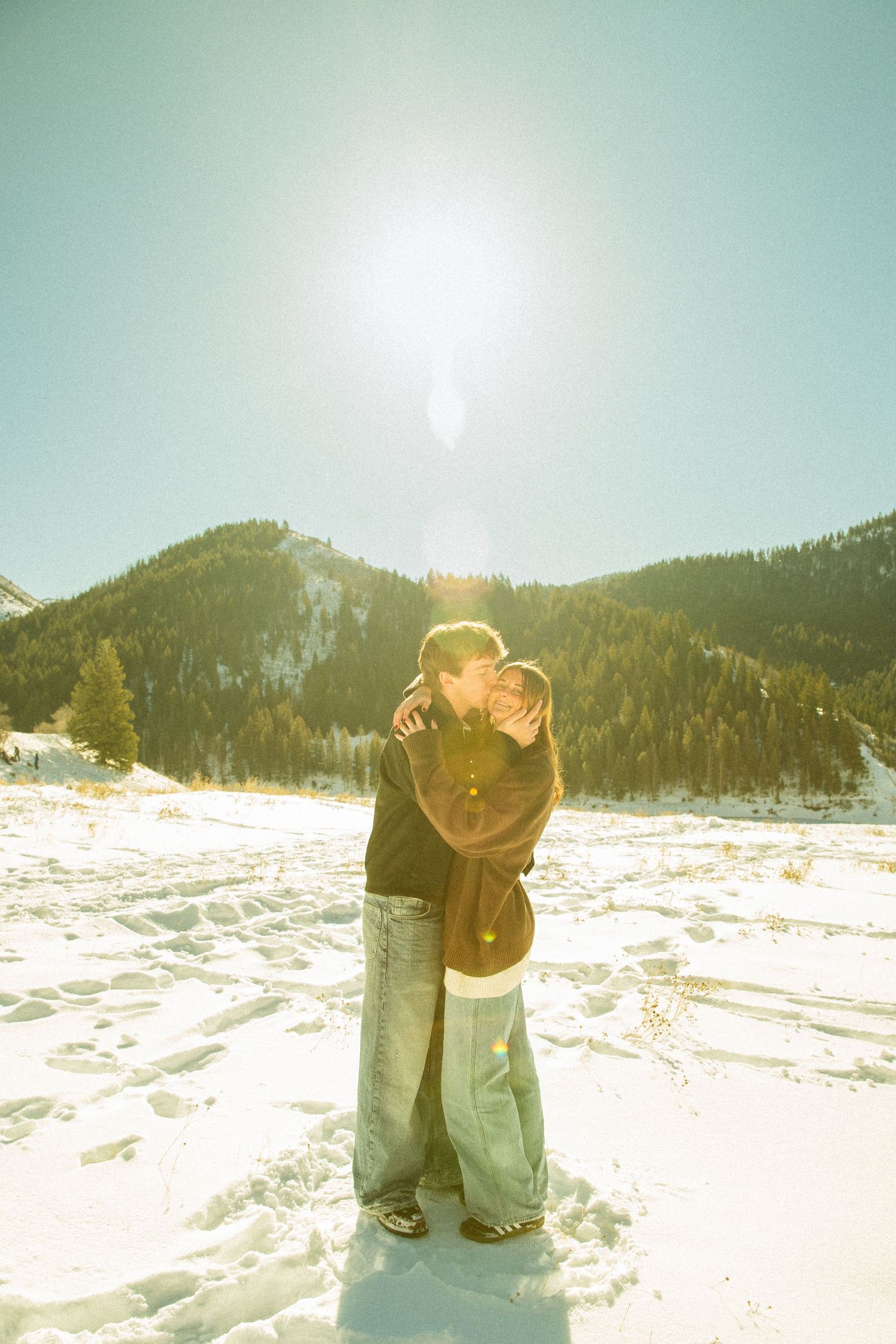 A romantic winter scene shows a couple embracing in the bright sunlight on a snowy mountain landscape.