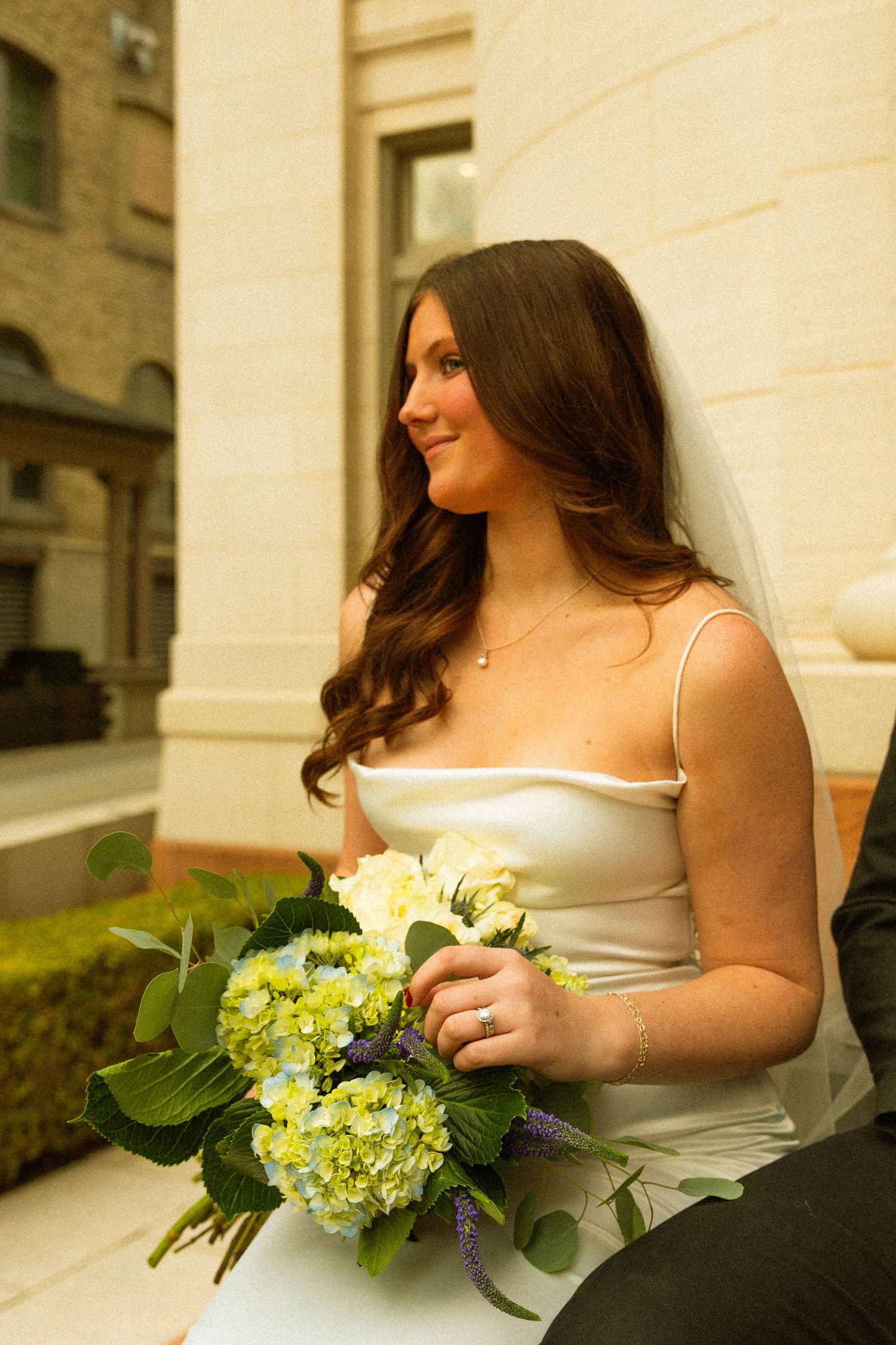 A bride in a white satin dress holds a bouquet of yellow hydrangeas while sitting outside a stone building.
