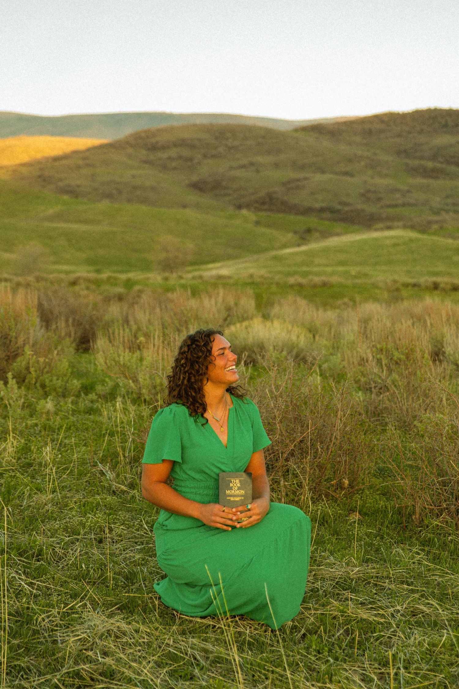 Woman in green dress sitting in grassy field, smiling, holding a jar, with rolling green hills in background.