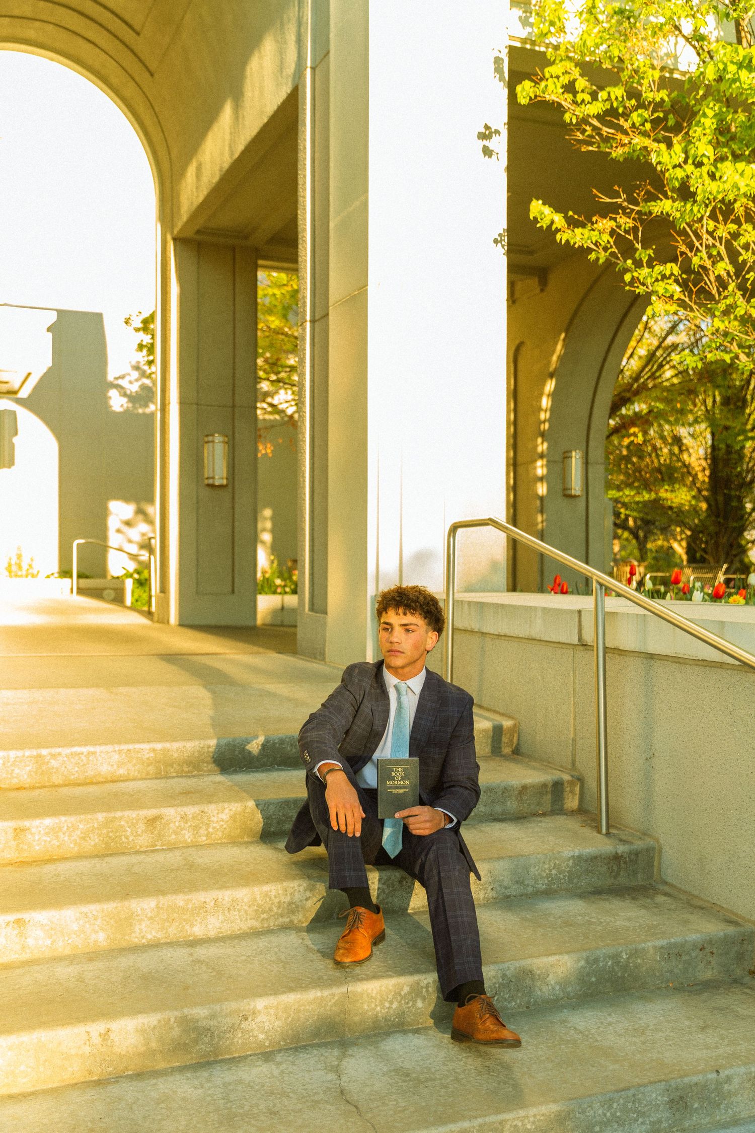Young man in dark suit holding a book sits on concrete steps outside an elegant building with arched columns.
