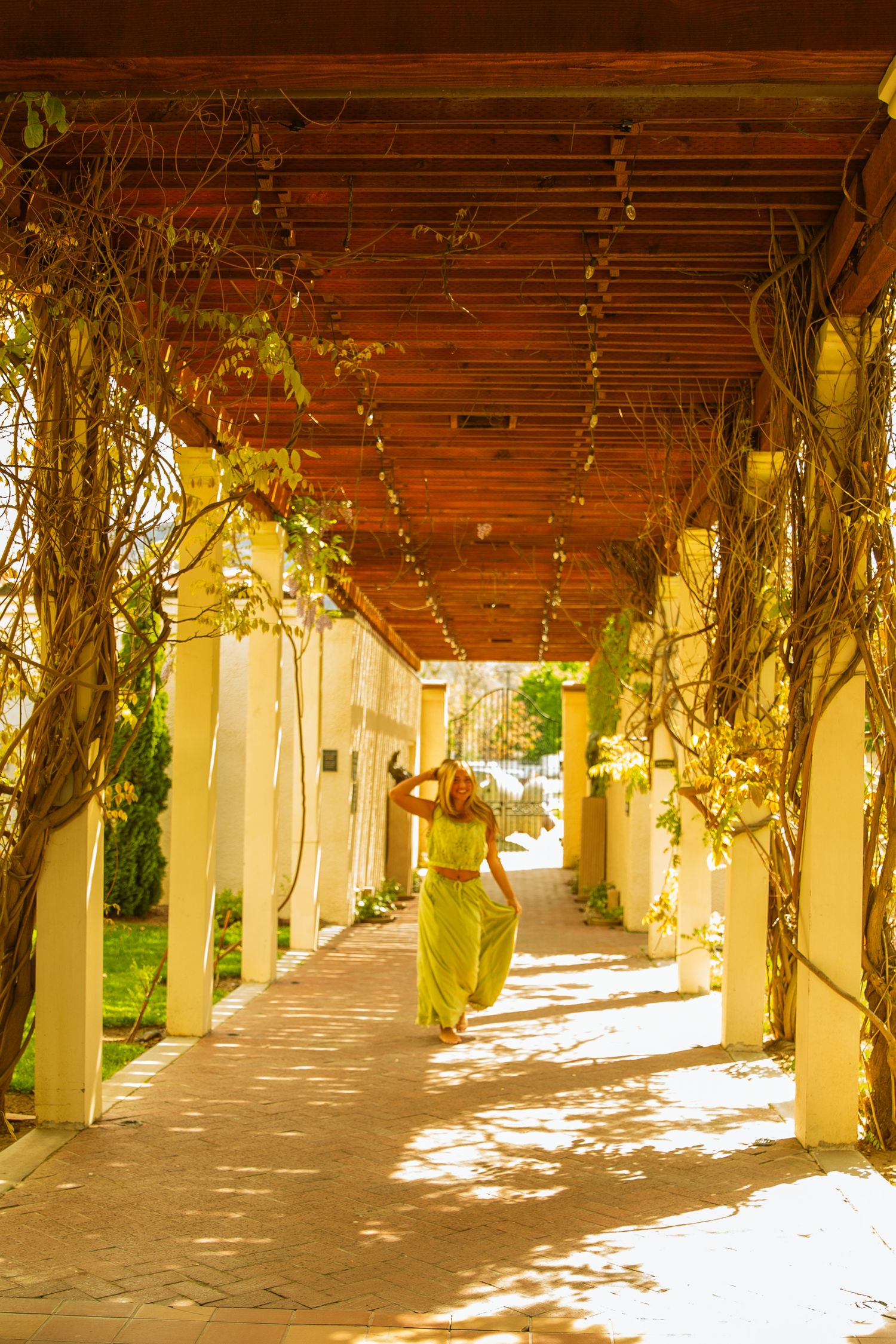Woman in yellow maxi dress walking through sun-dappled pergola corridor with climbing vines and white columns.