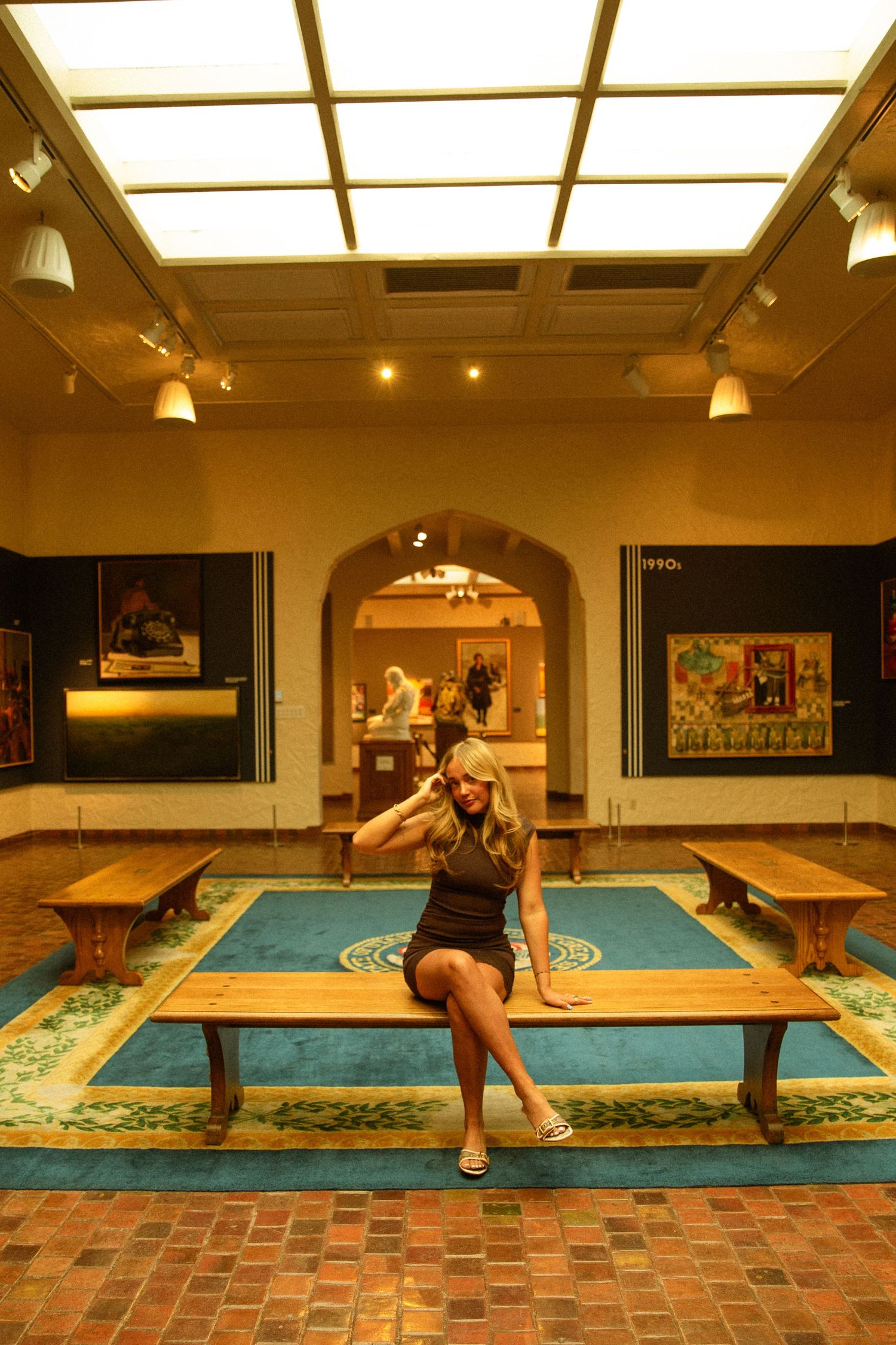 Blonde woman sitting on wooden bench inside an art museum gallery with blue rug and paintings on walls.