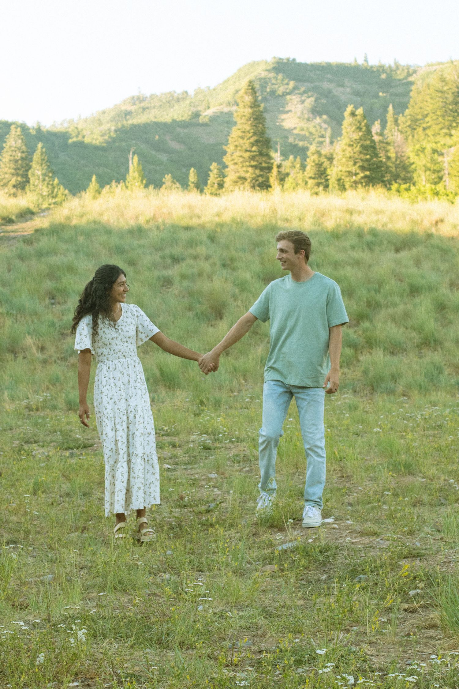 A couple holds hands while walking together in a grassy field at sunset with mountains in the background.