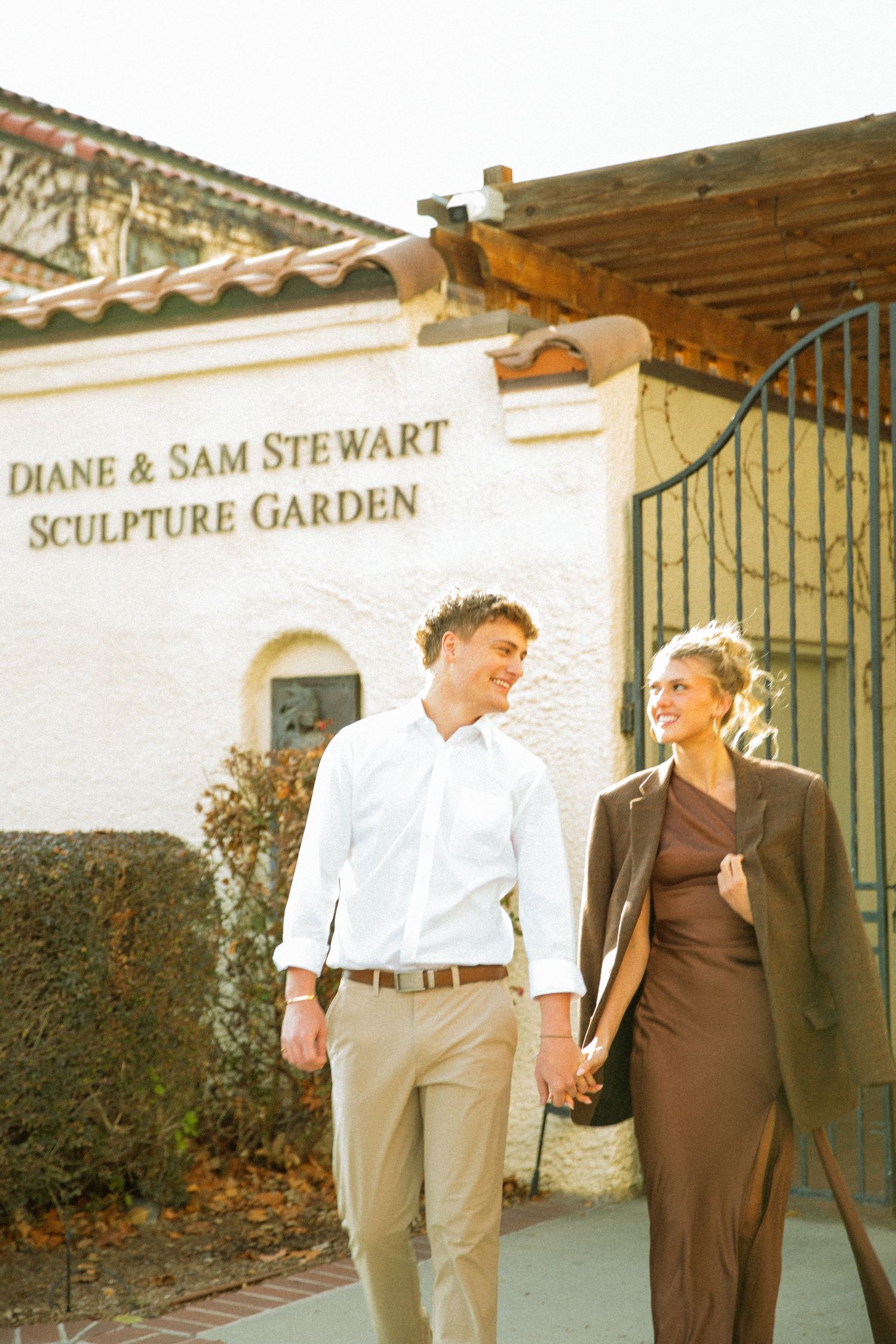 A couple in brown and white outfits walk and pose together in front of a garden wall with Spanish-style architecture.