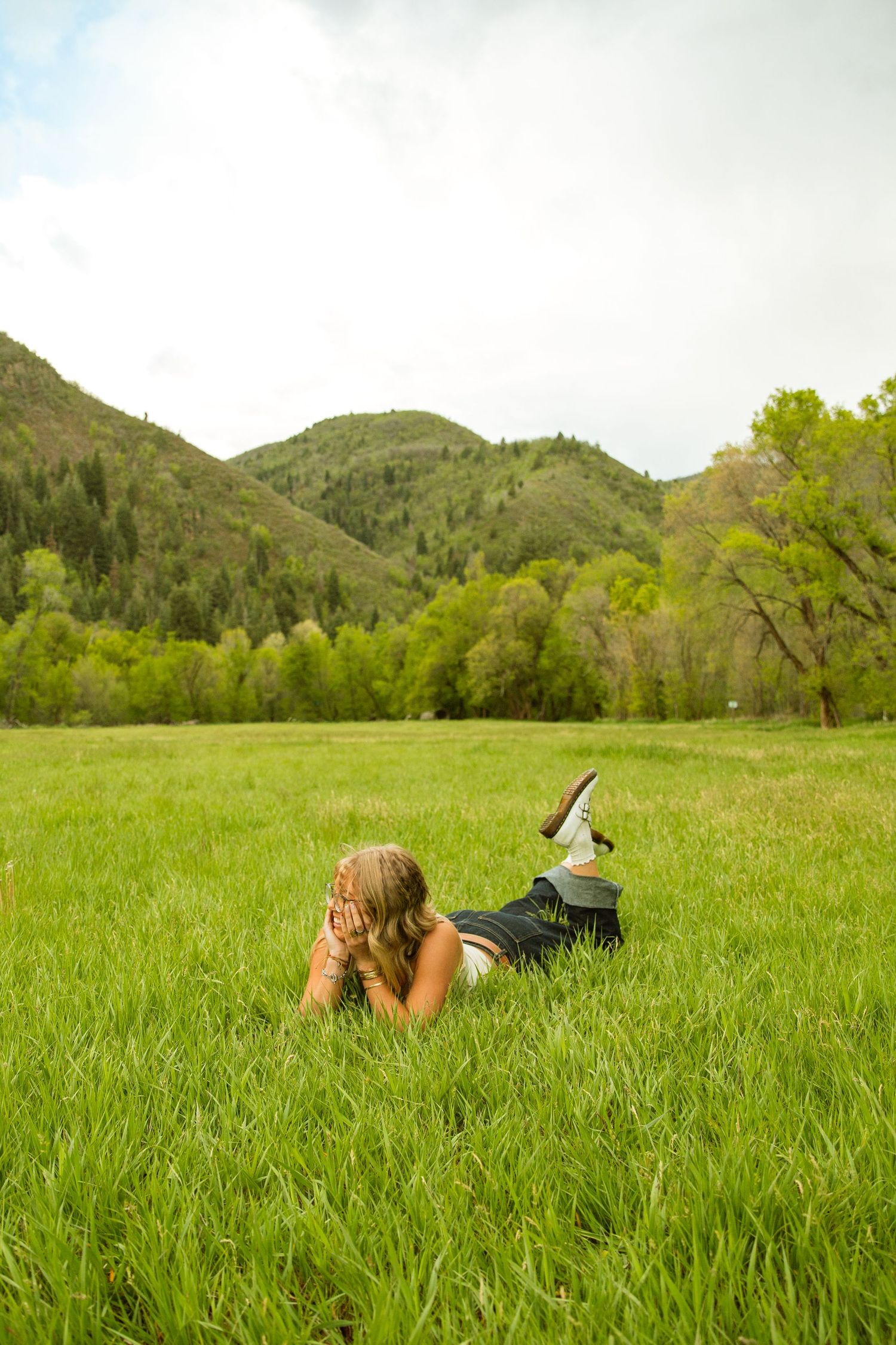 Woman lying in lush green meadow with mountains and forest in background on a cloudy day.