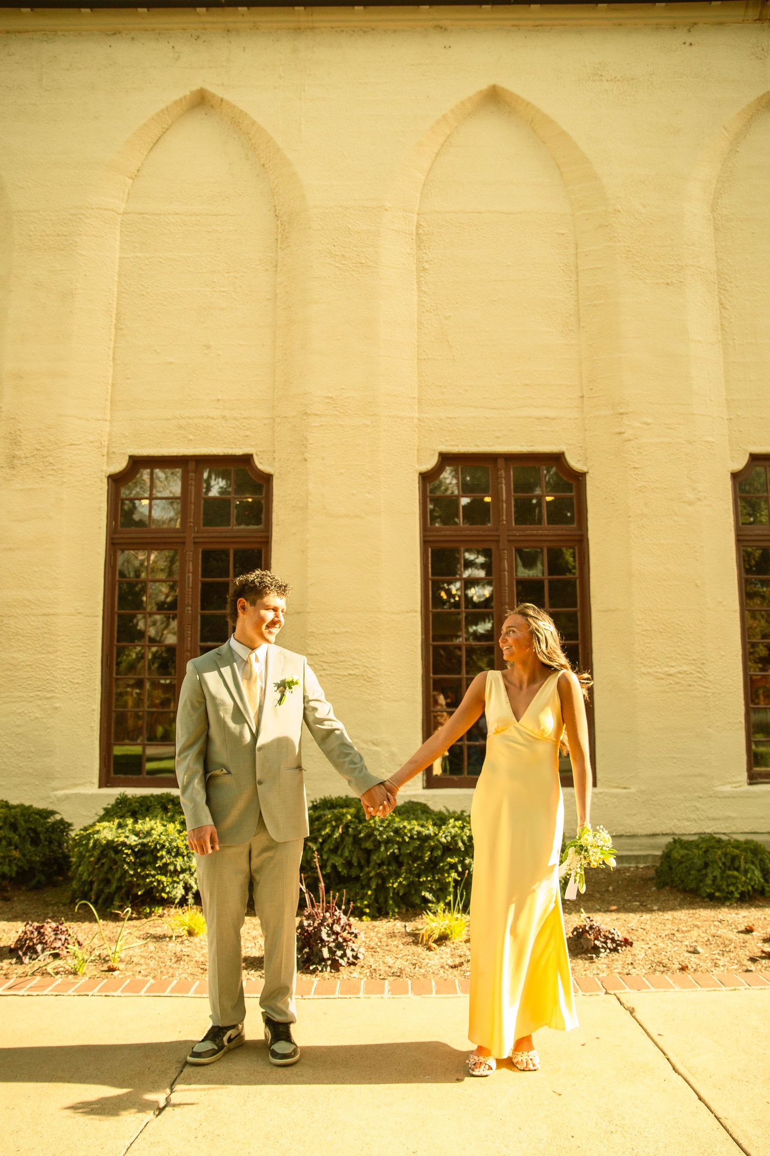 A couple in formal attire holds hands outside a white brick building with arched windows on a sunny day.