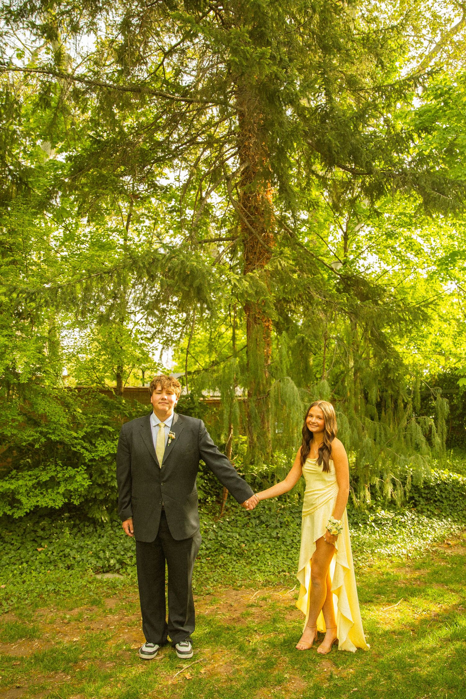 A young couple dressed in formal prom attire holds hands outdoors amid lush green trees.