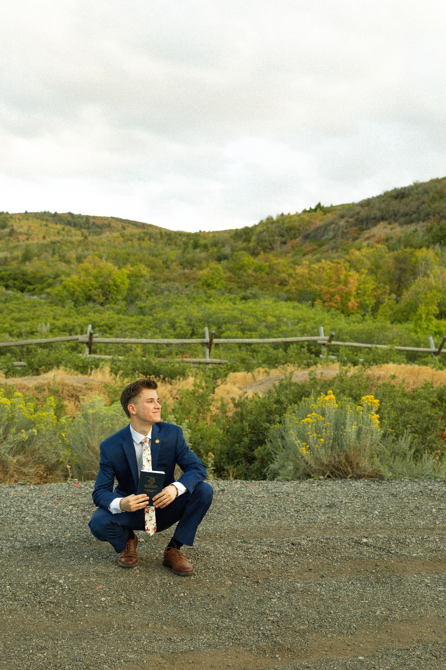 Person in navy suit crouches down on gravel path with scenic mountain landscape and autumn foliage in background.