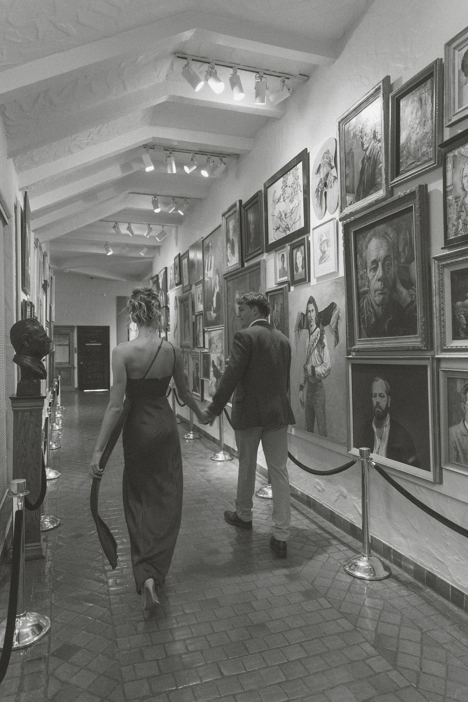 Museum visitors walk through a gallery corridor with framed portraits and artwork displayed on white walls under vaulted lighting.