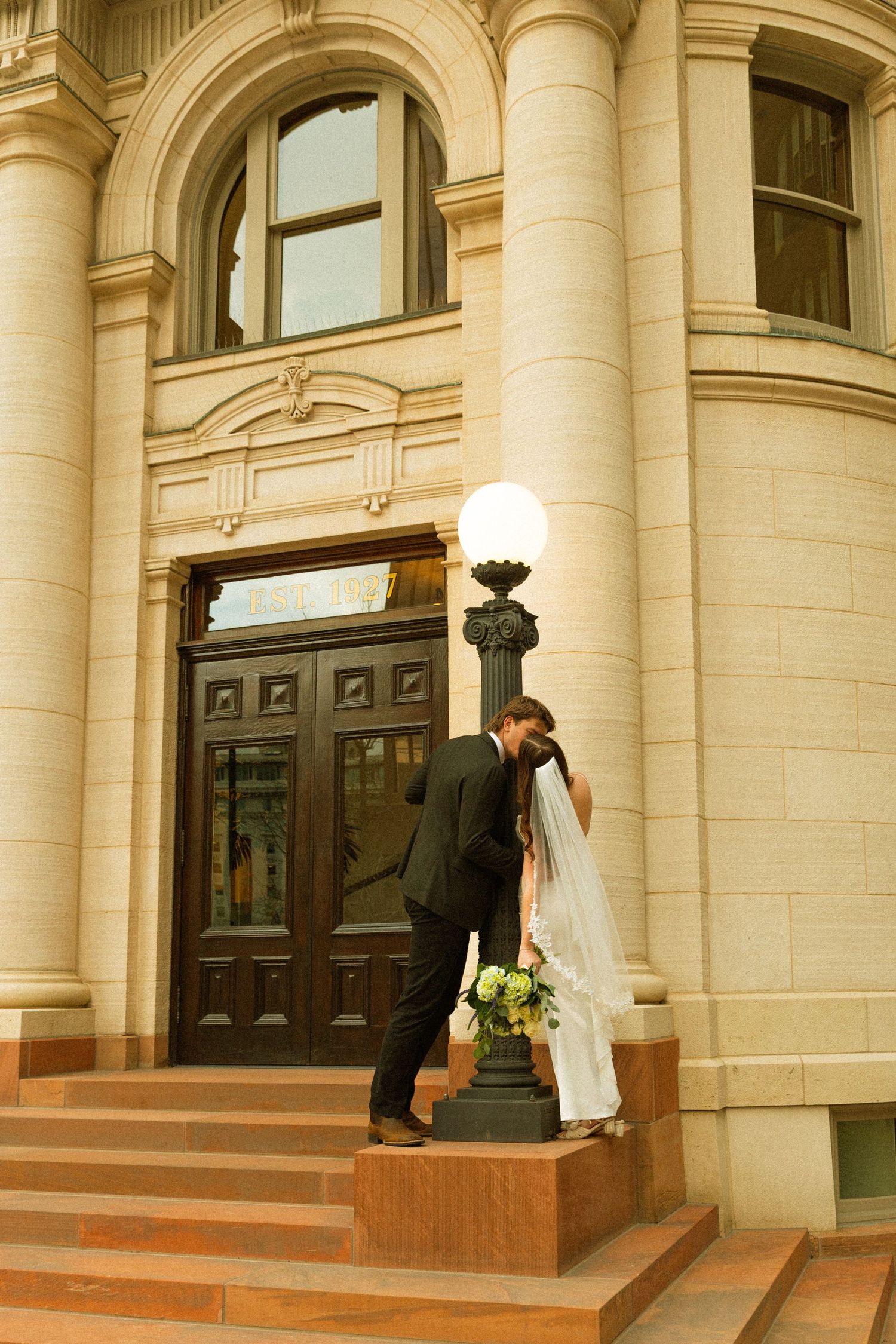 A bride and groom share an intimate moment on the steps of a grand building with classical stone columns.