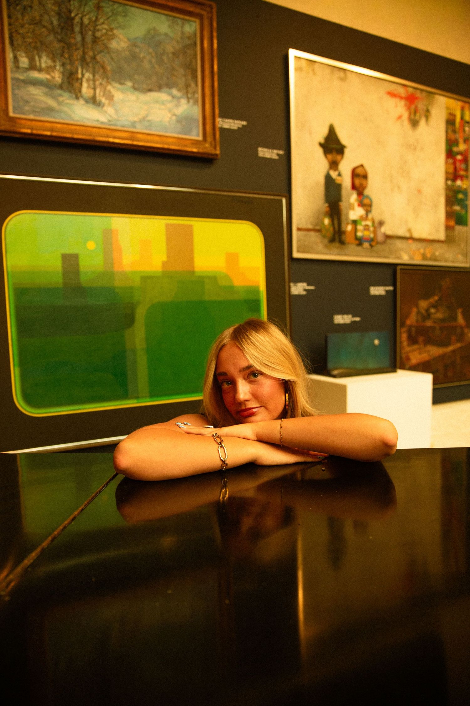 Blonde woman resting chin on arms at a museum display case surrounded by colorful paintings on a dark wall.
