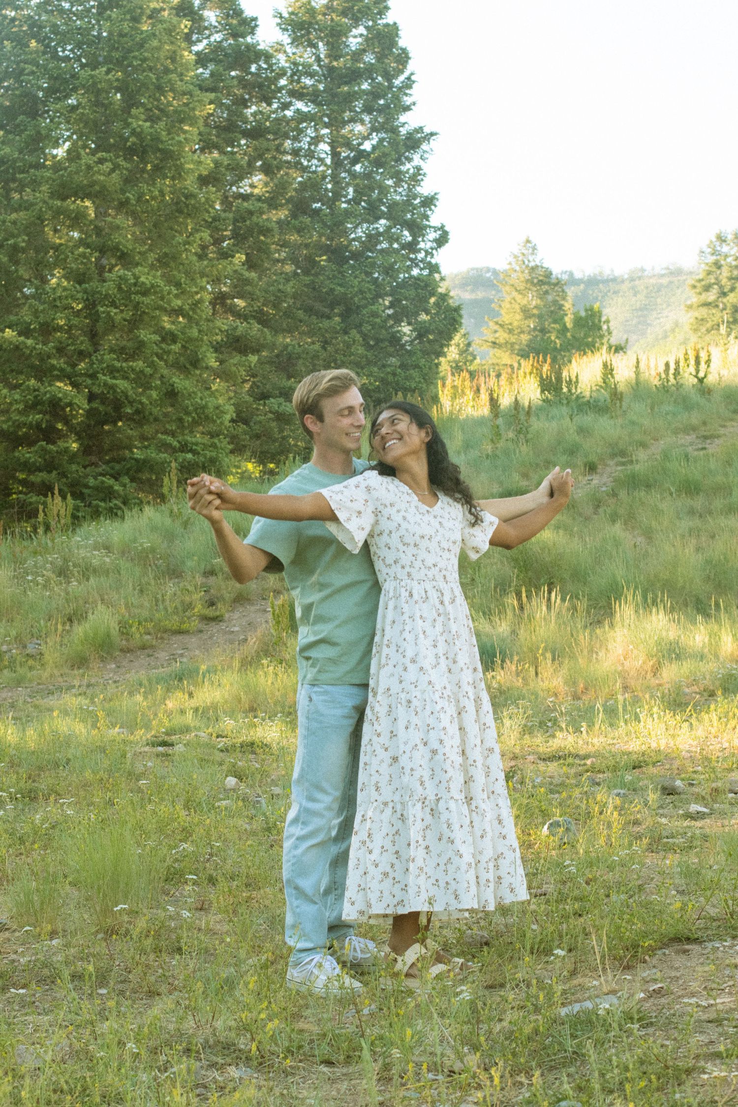 A couple dances together in a sunlit meadow surrounded by evergreen trees during golden hour.