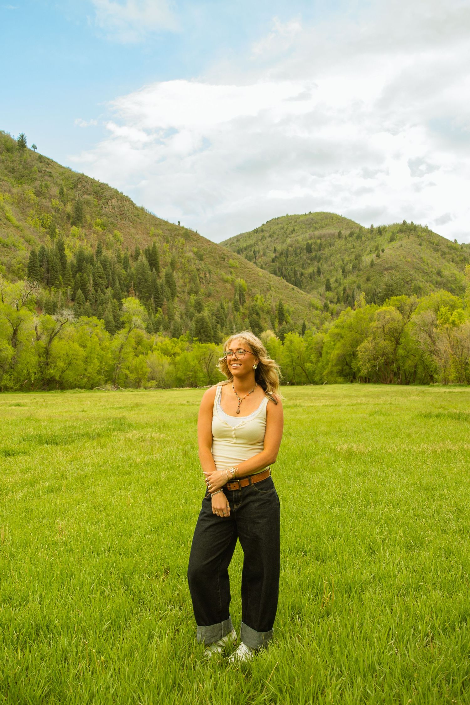 Happy blonde woman in white tank top and black jeans posing in green field with scenic mountain views.