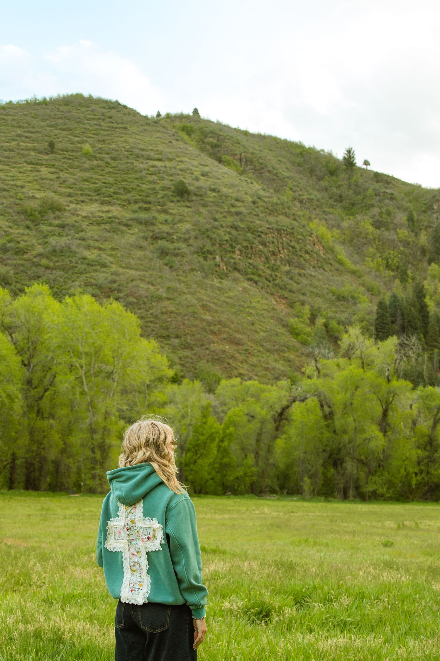 Blonde girl in green cross hoodie stands in lush meadow with tree-covered hill backdrop.