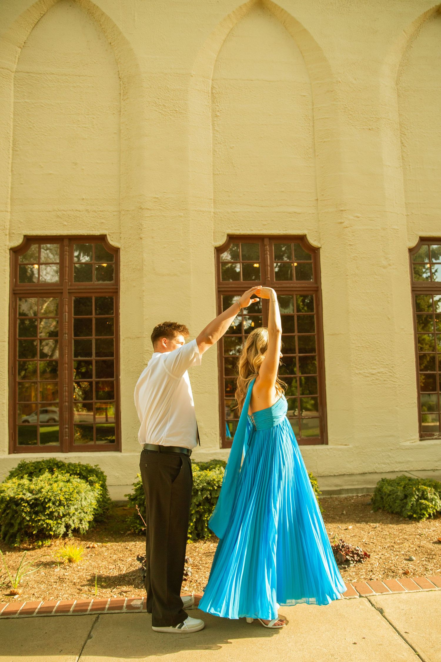 Couple dancing outdoors in formal wear, woman in blue pleated gown, man in white shirt, beside elegant arched building.