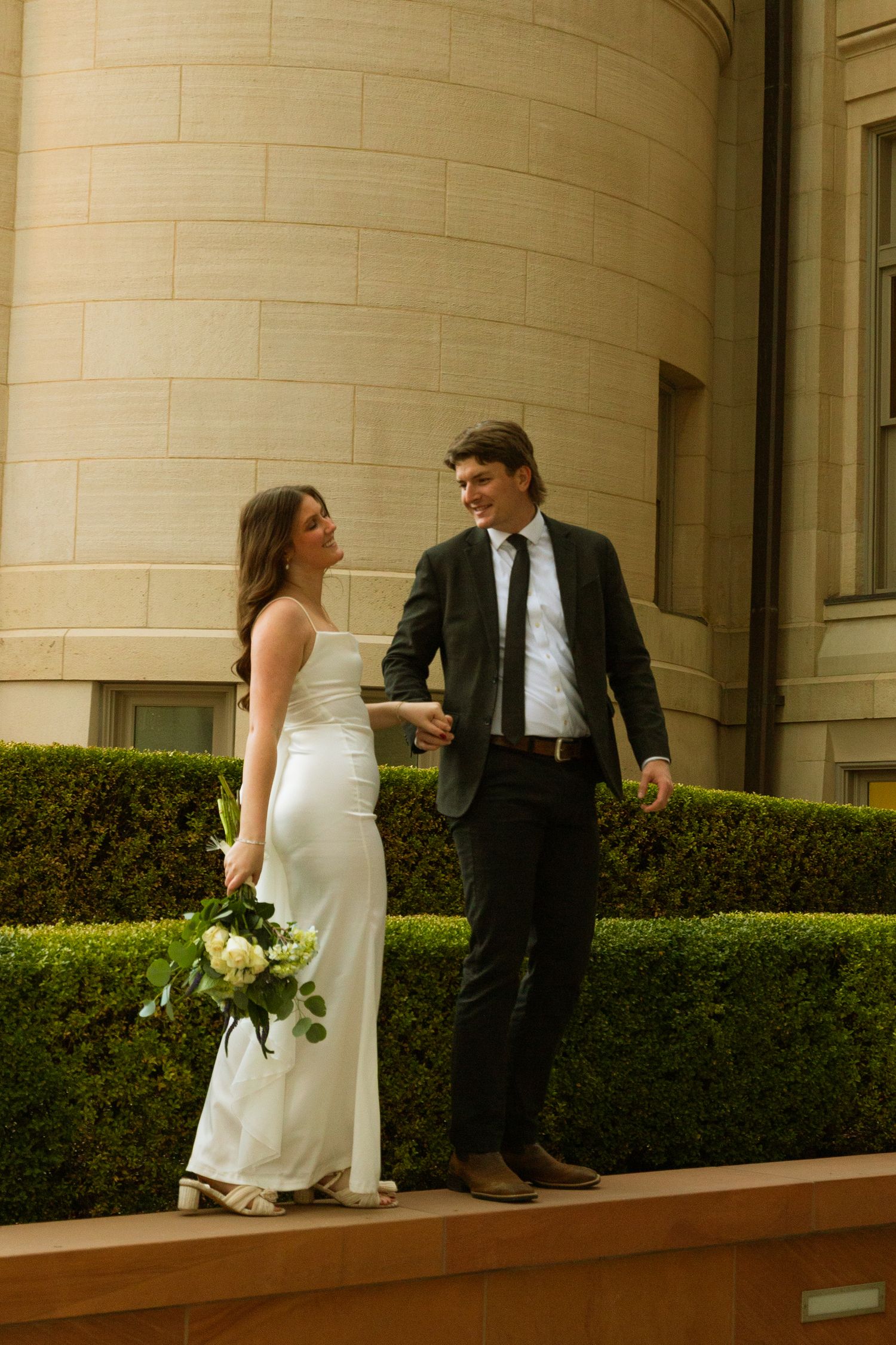 A couple in wedding attire stands together on a wooden deck against a curved beige wall with manicured hedges.