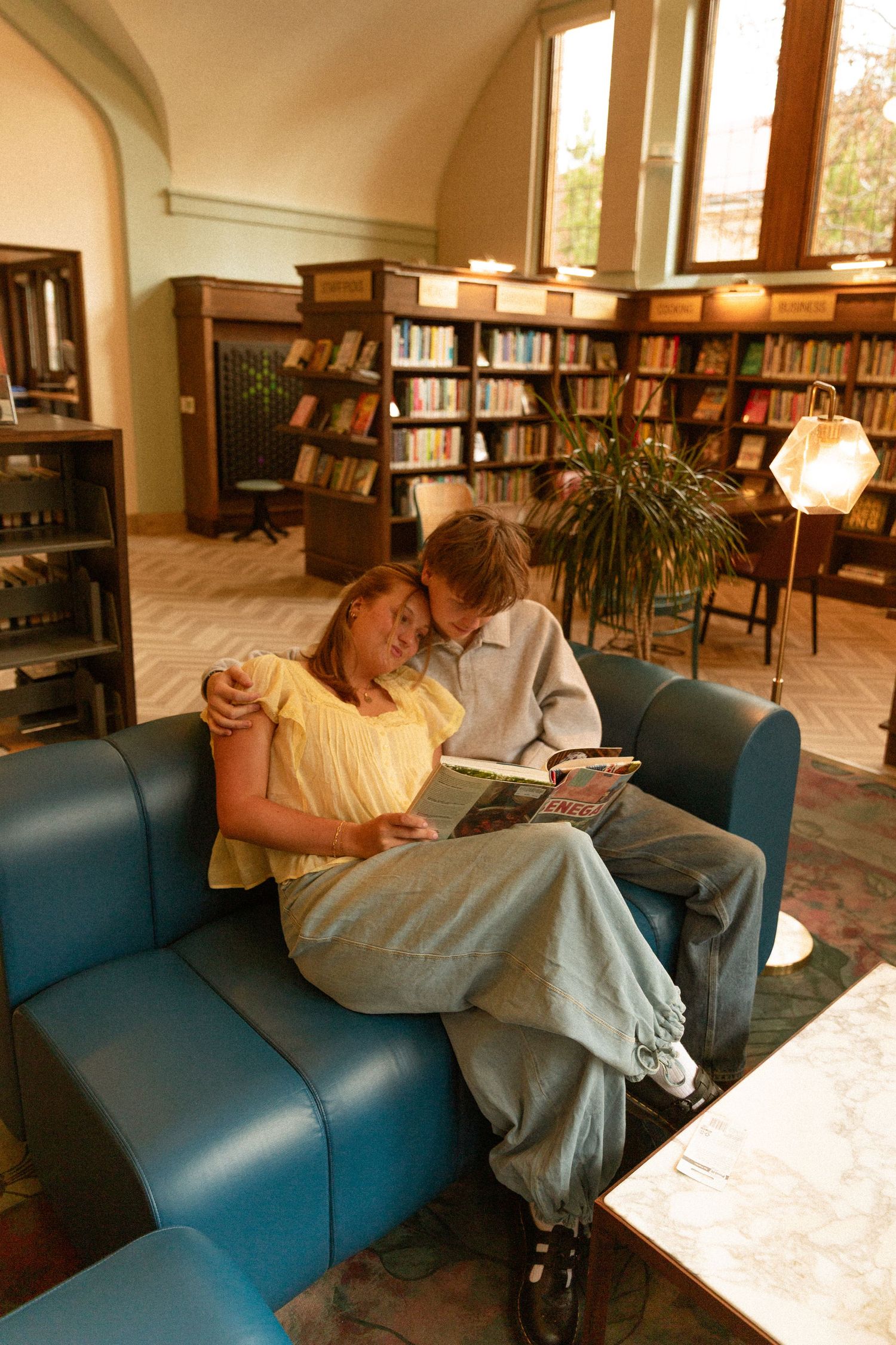 Students resting and studying on blue couches in a library with bookshelves visible in the background.