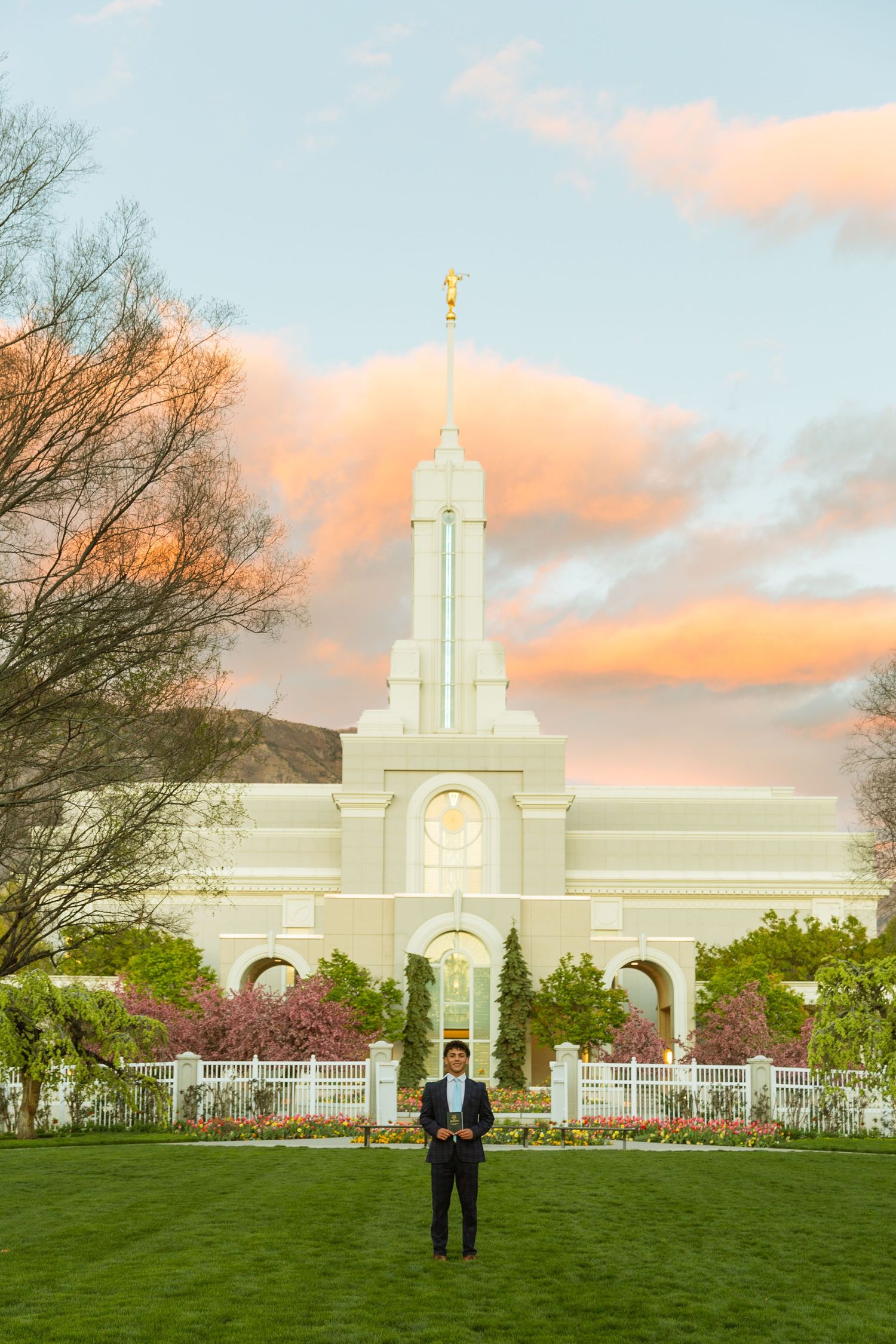 Man stands on green lawn before white LDS temple with golden spire under a pastel sunset sky with pink clouds.
