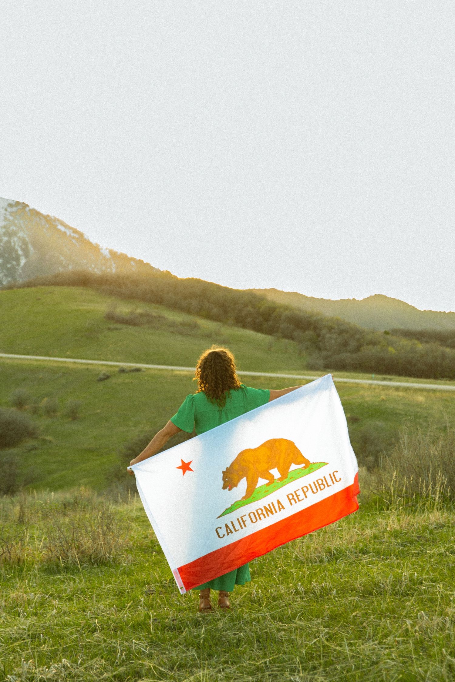 Person holding California Republic state flag in green meadow with rolling hills and mountains in background.