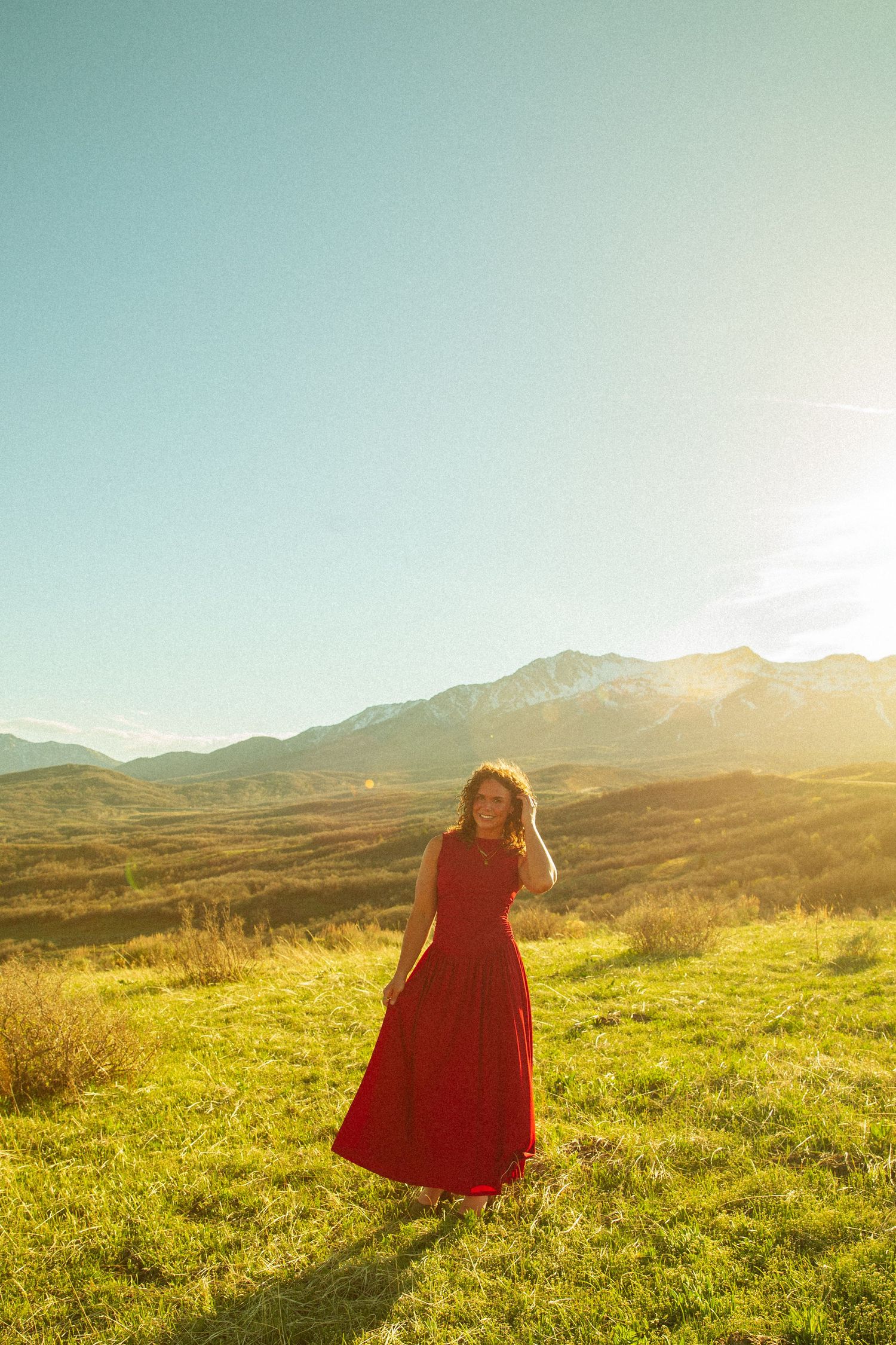 Woman in red maxi dress standing in sunlit green meadow with mountain backdrop on a clear day.