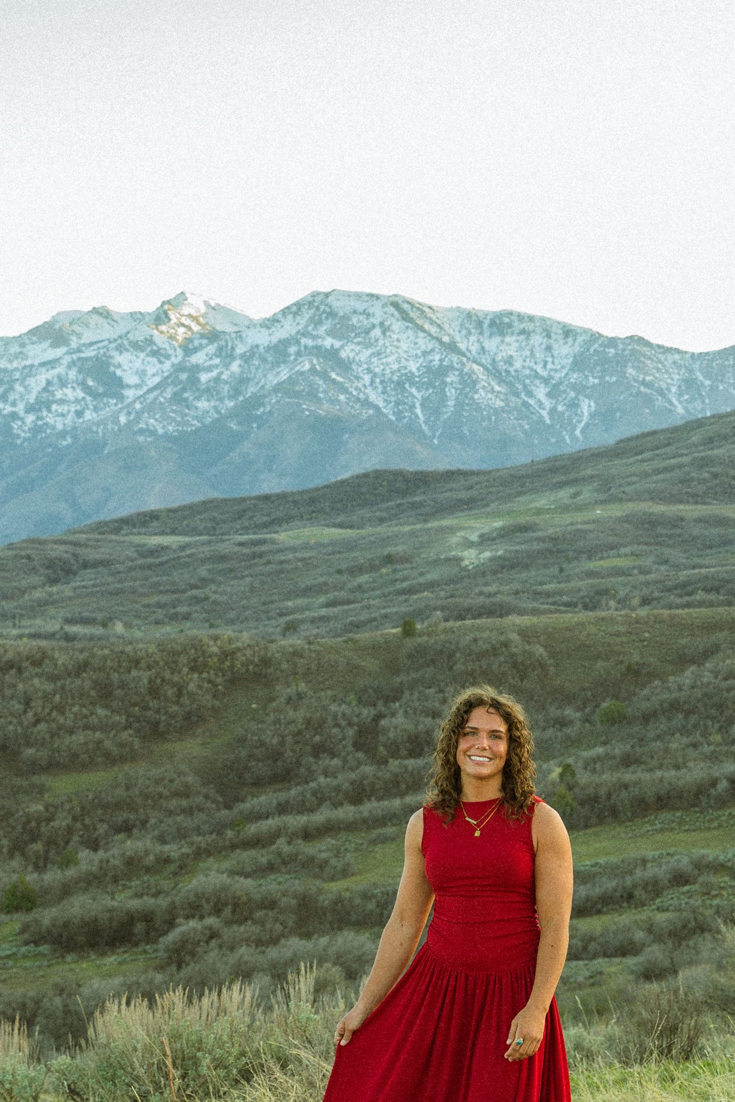 A smiling young woman in a red dress stands before a stunning snow-capped mountain and green valley landscape.