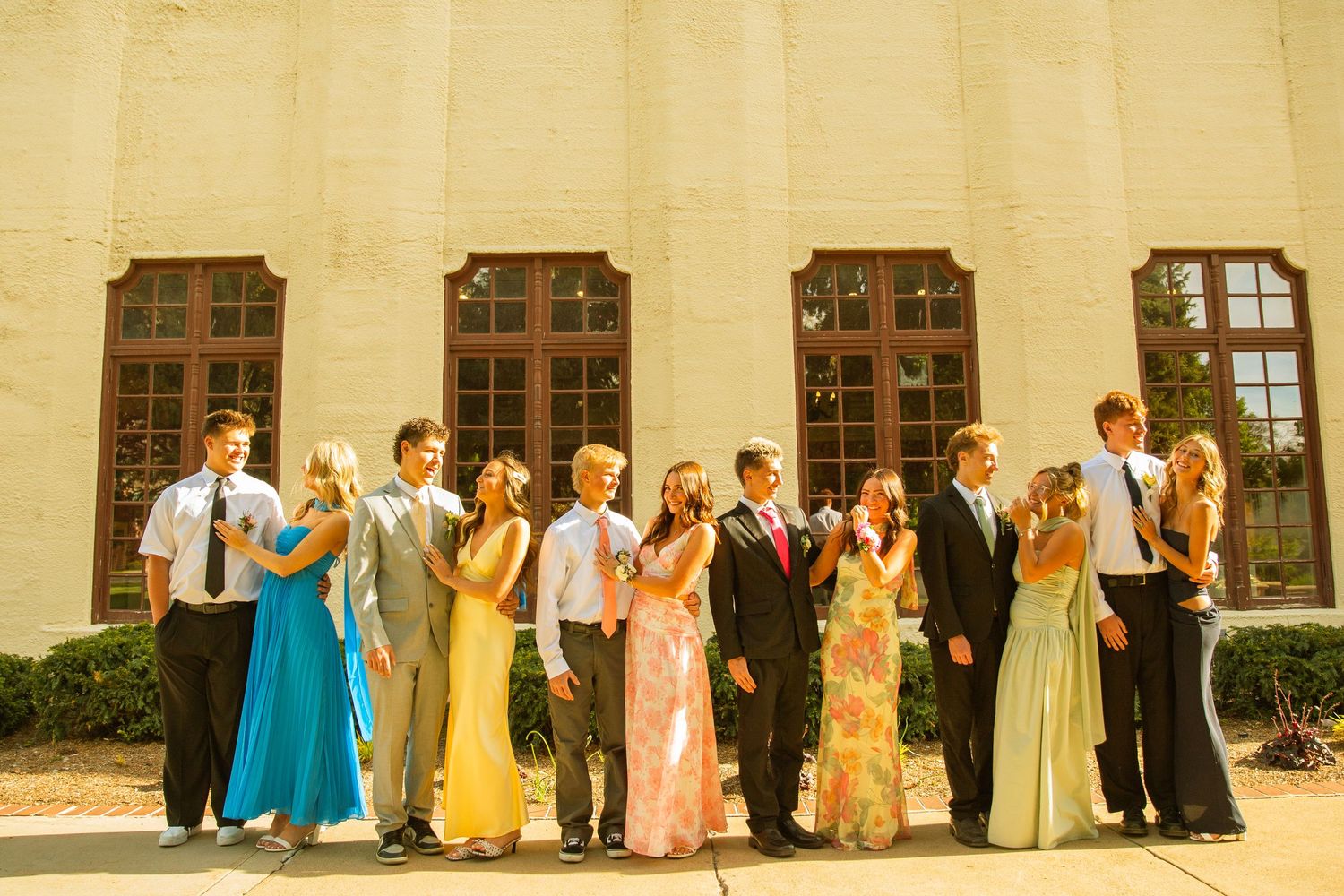 Group of teens in formal prom attire posing outside a white brick building on a sunny day.