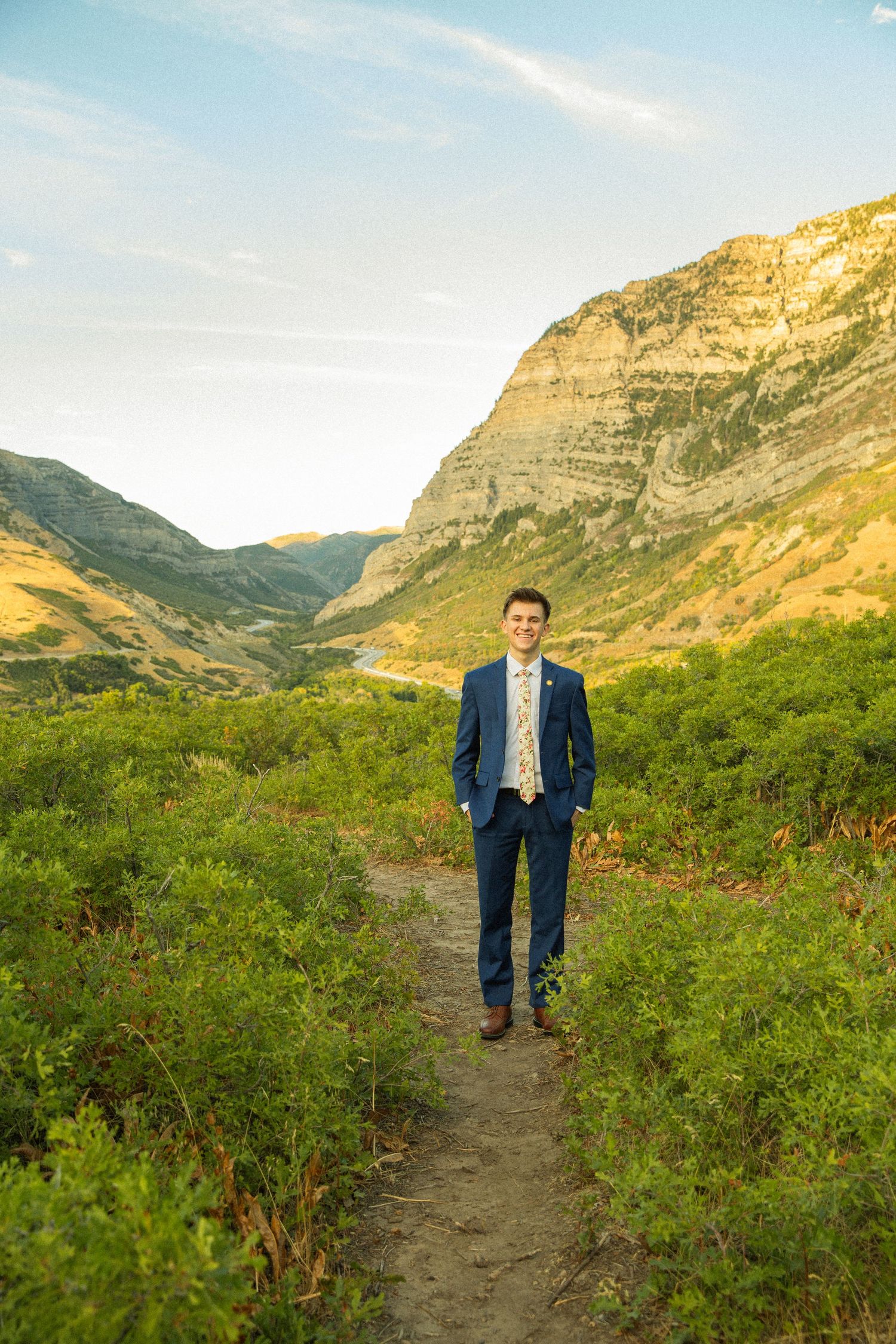A dirt hiking trail winds through green shrubs with dramatic mountain cliffs and golden sunlight in Utah's wilderness.