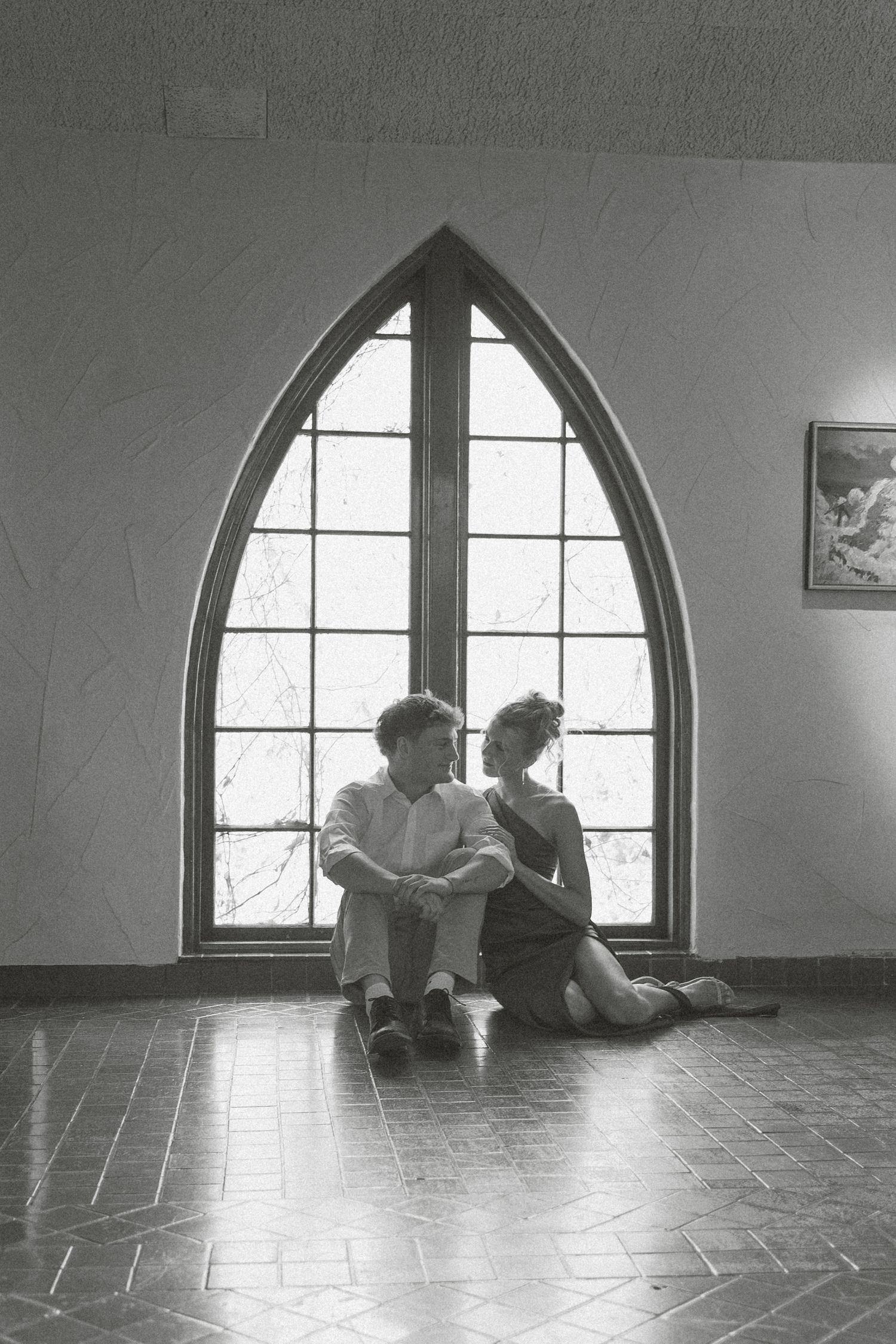 Two figures sit silently by an arched window in a minimalist room with tiled floors.