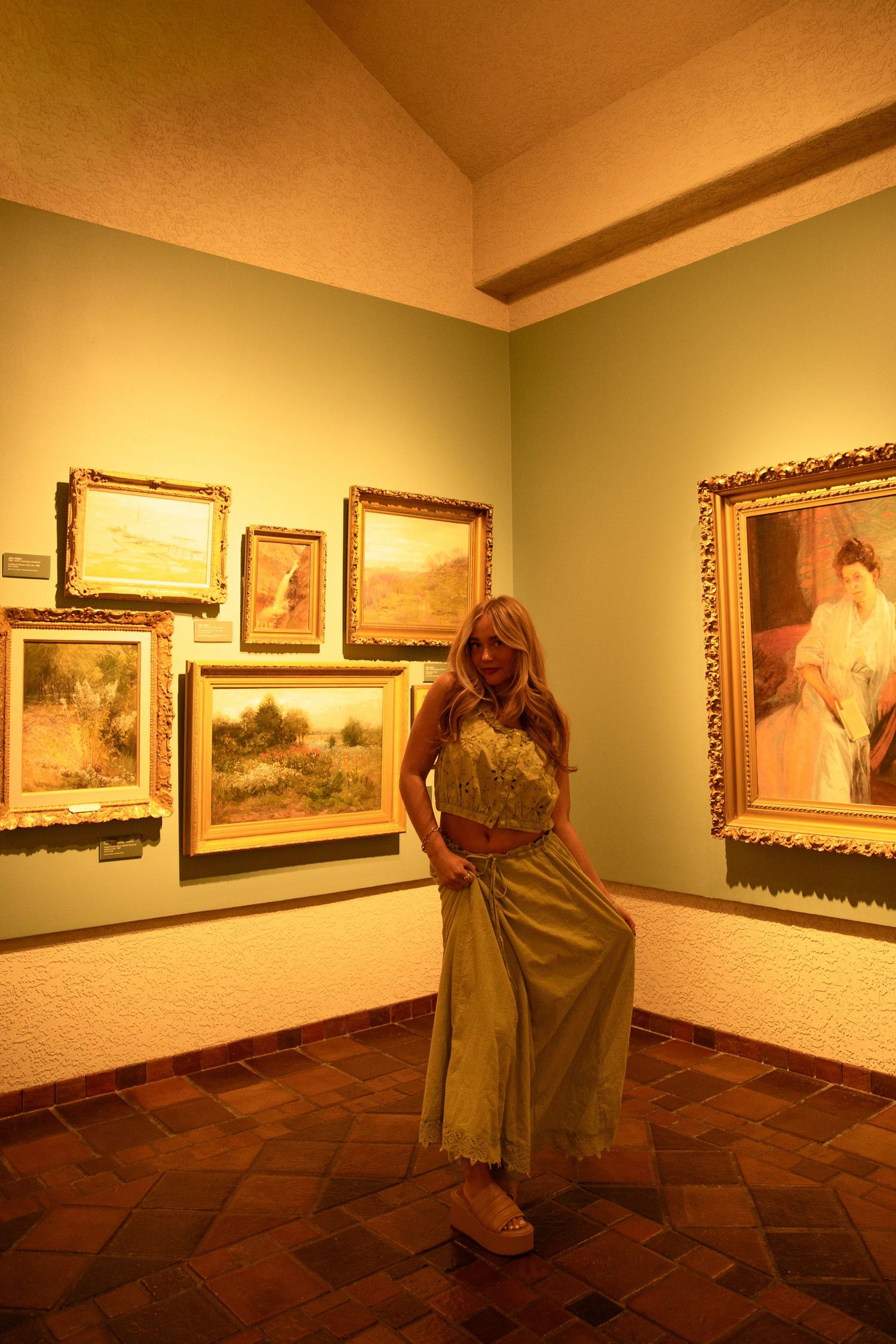 Woman in green outfit posing in art gallery corner surrounded by gold-framed paintings on sage green walls.