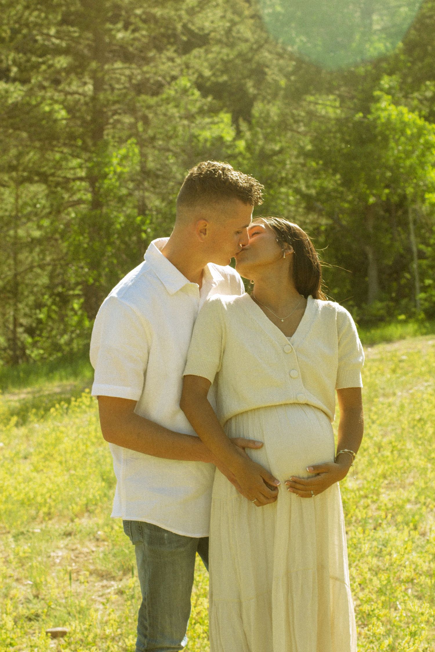 A couple in white outfits share tender moments together in a sunlit meadow with trees in the background.