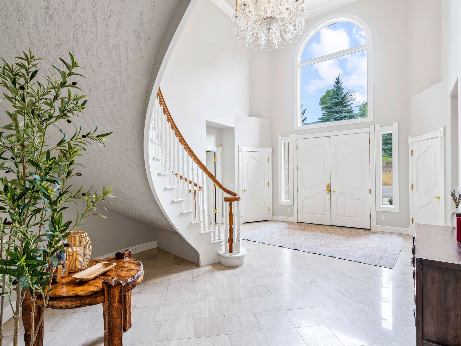 Grand foyer with curved staircase, chandelier, arched window, white double doors, and marble tile floors.