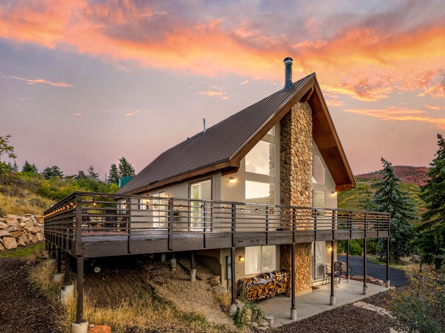 Rustic A-frame cabin with stone exterior, large deck with metal railings, and vibrant orange sunset backdrop.