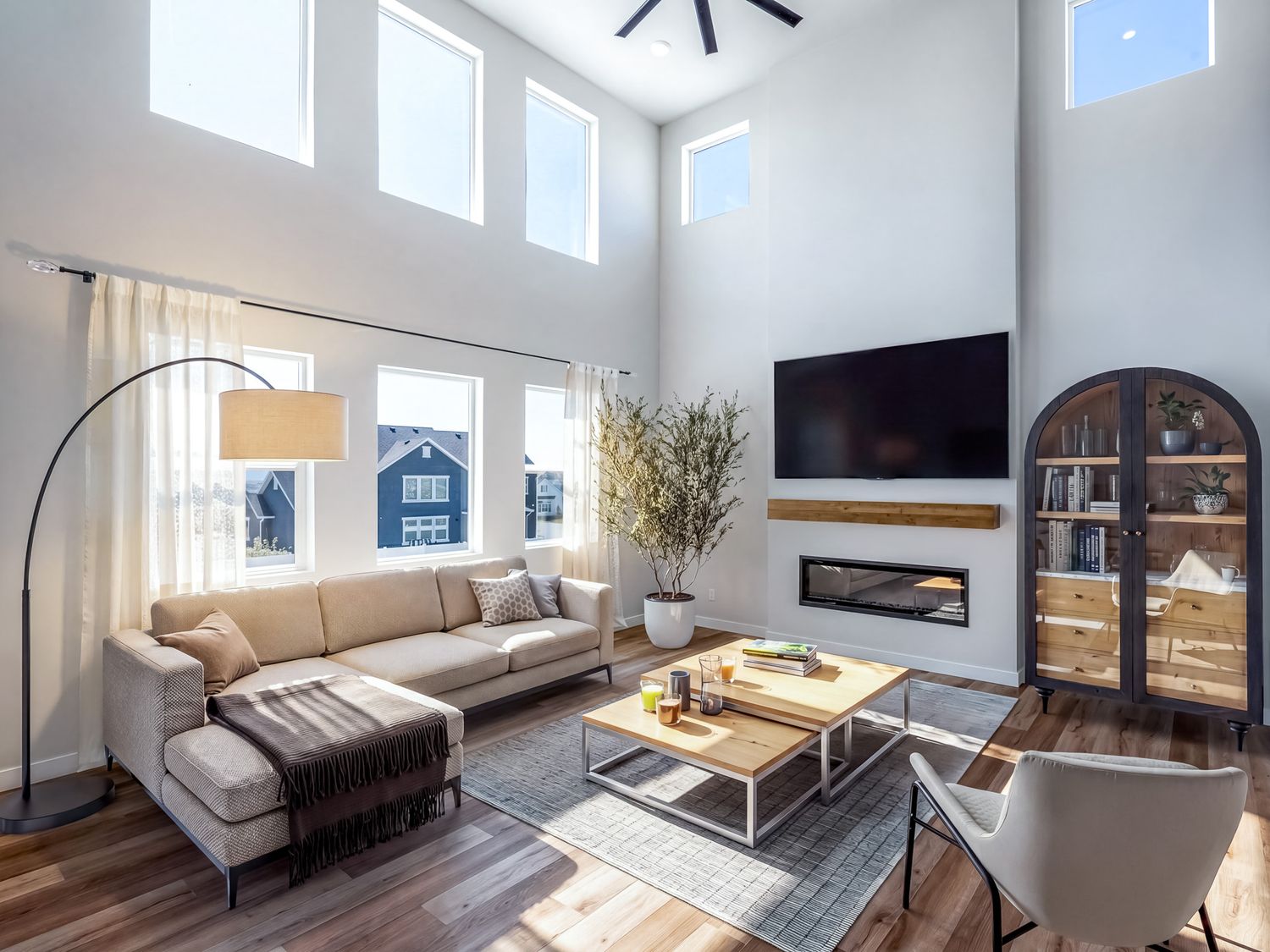 Bright modern living room with high ceilings, large windows, beige sectional sofa, wood floors, and wall-mounted TV above fireplace.