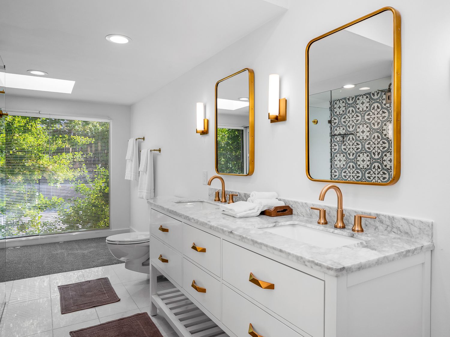 Bright white bathroom with gold-framed mirrors, rose gold fixtures, marble countertop, and patterned shower tile.