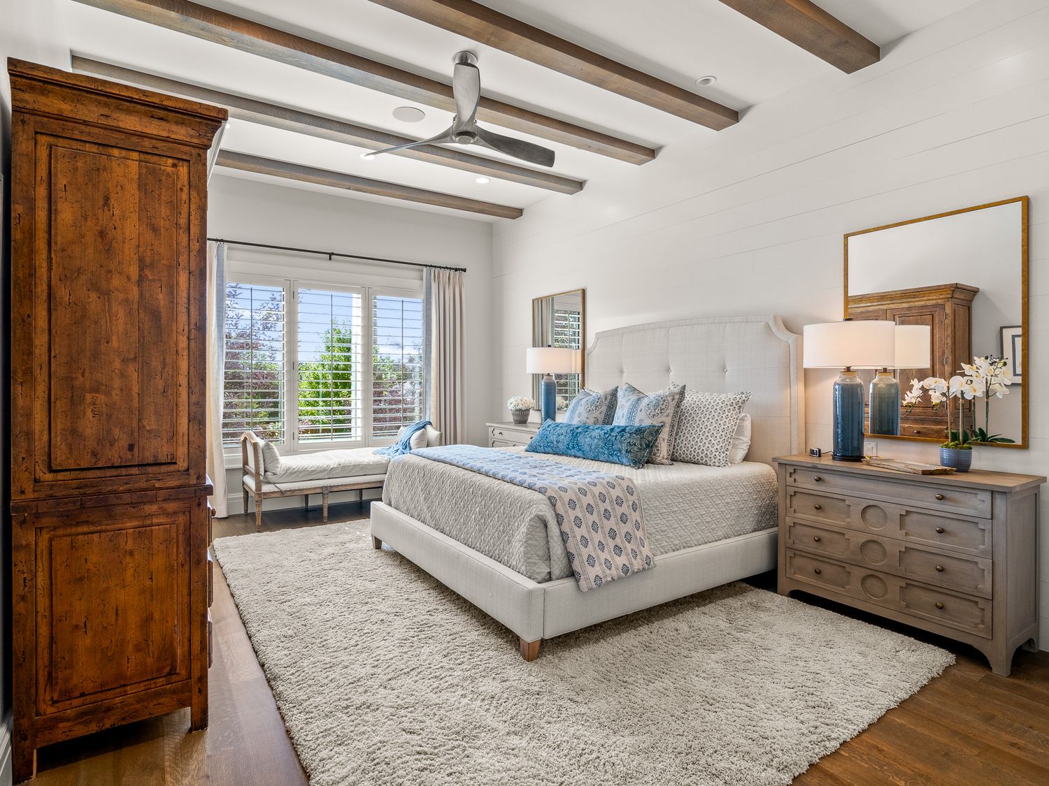 Spacious master bedroom with exposed wood beam ceiling, upholstered bed, and hardwood floors with plush white area rug.