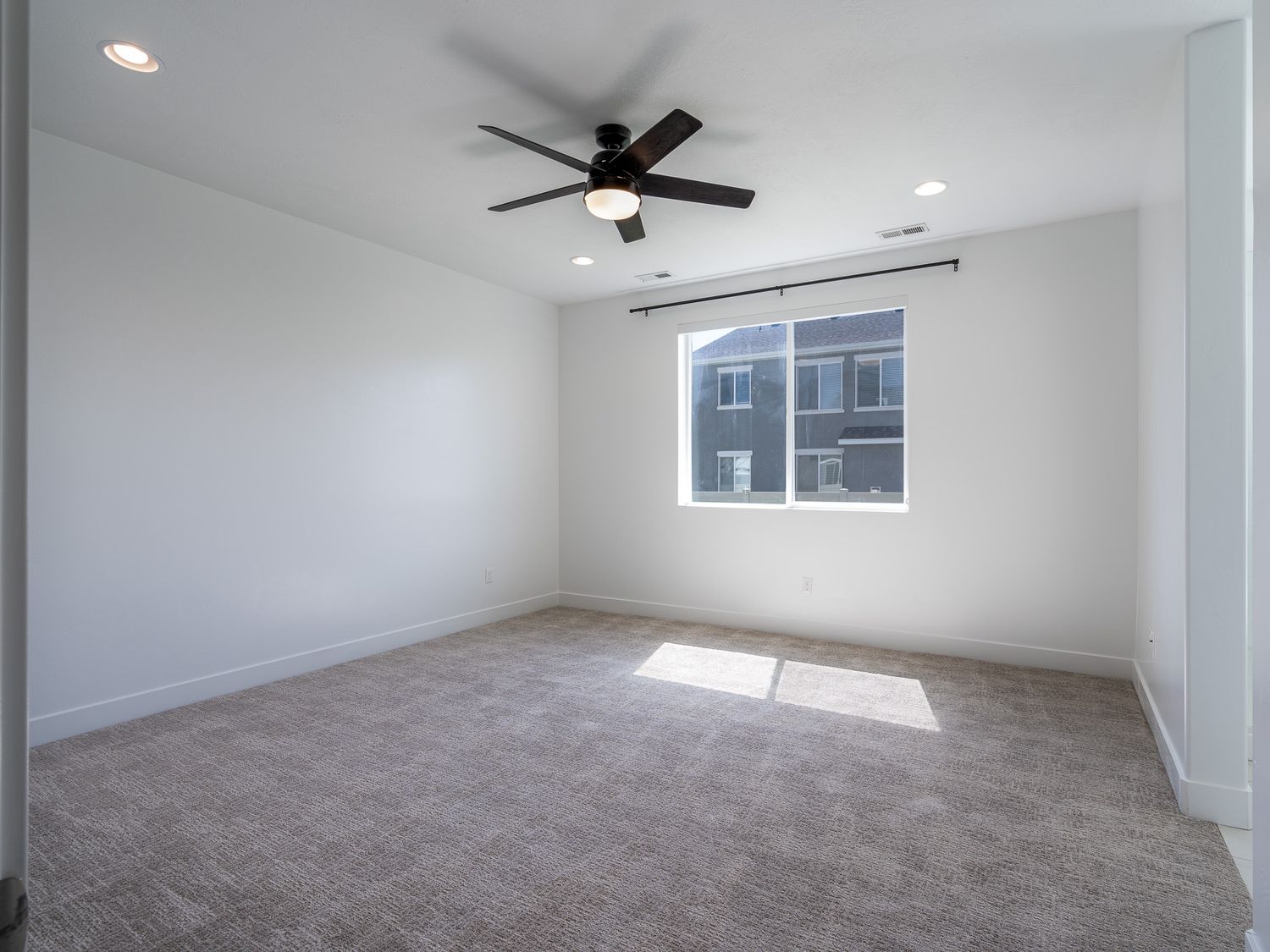 Minimalist empty bedroom with gray carpet, ceiling fan, recessed lighting, and a single window with neighborhood view.