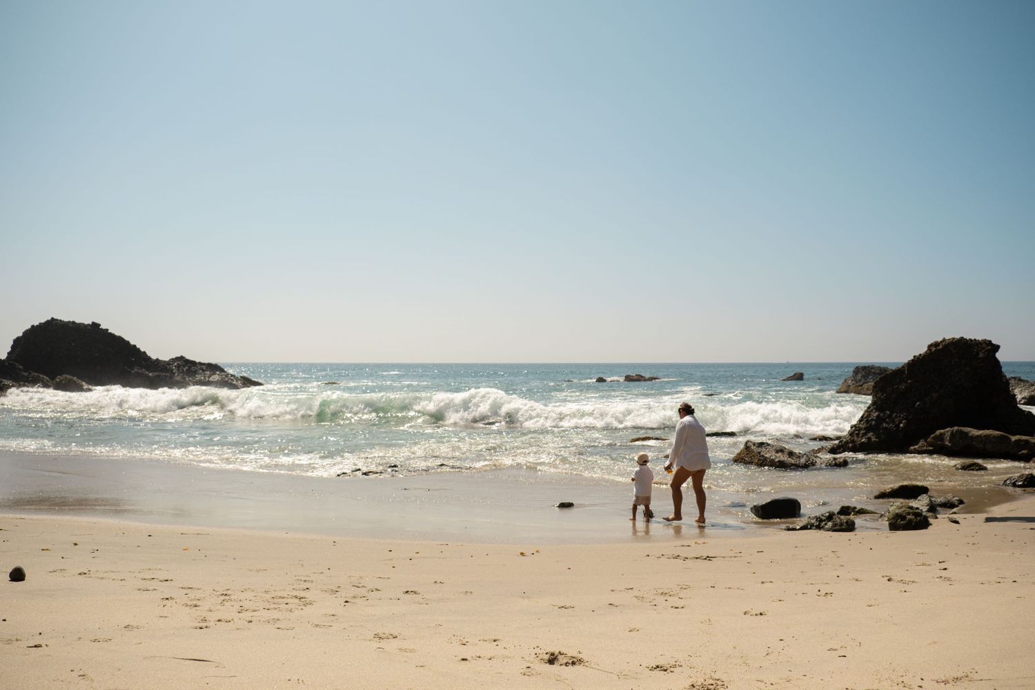 Waves crash against rocky outcrops along a scenic beach coastline on a sunny day.